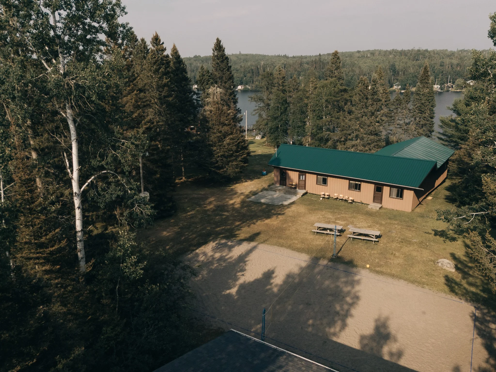 A house with a green metal roof surrounded by trees and a lake in the background under a cloudy sky.