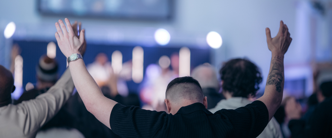 Person with short hair and tattoos raising hands in a crowd at an indoor concert or event.