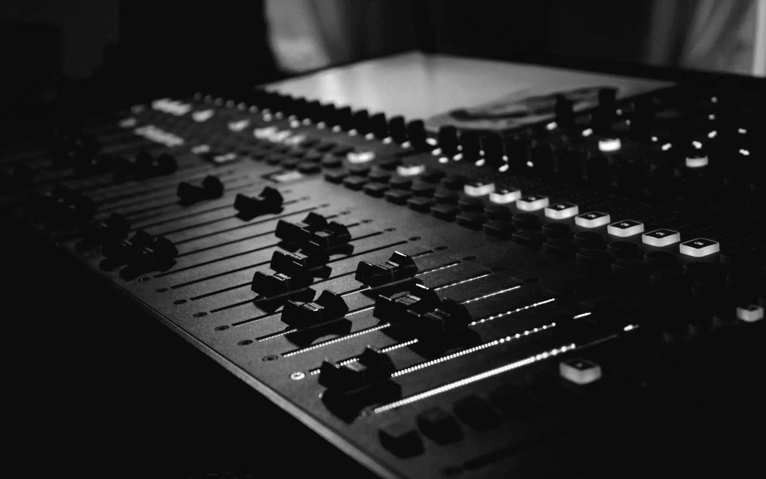 Close-up of an audio mixing console with illuminated faders and knobs in black and white.