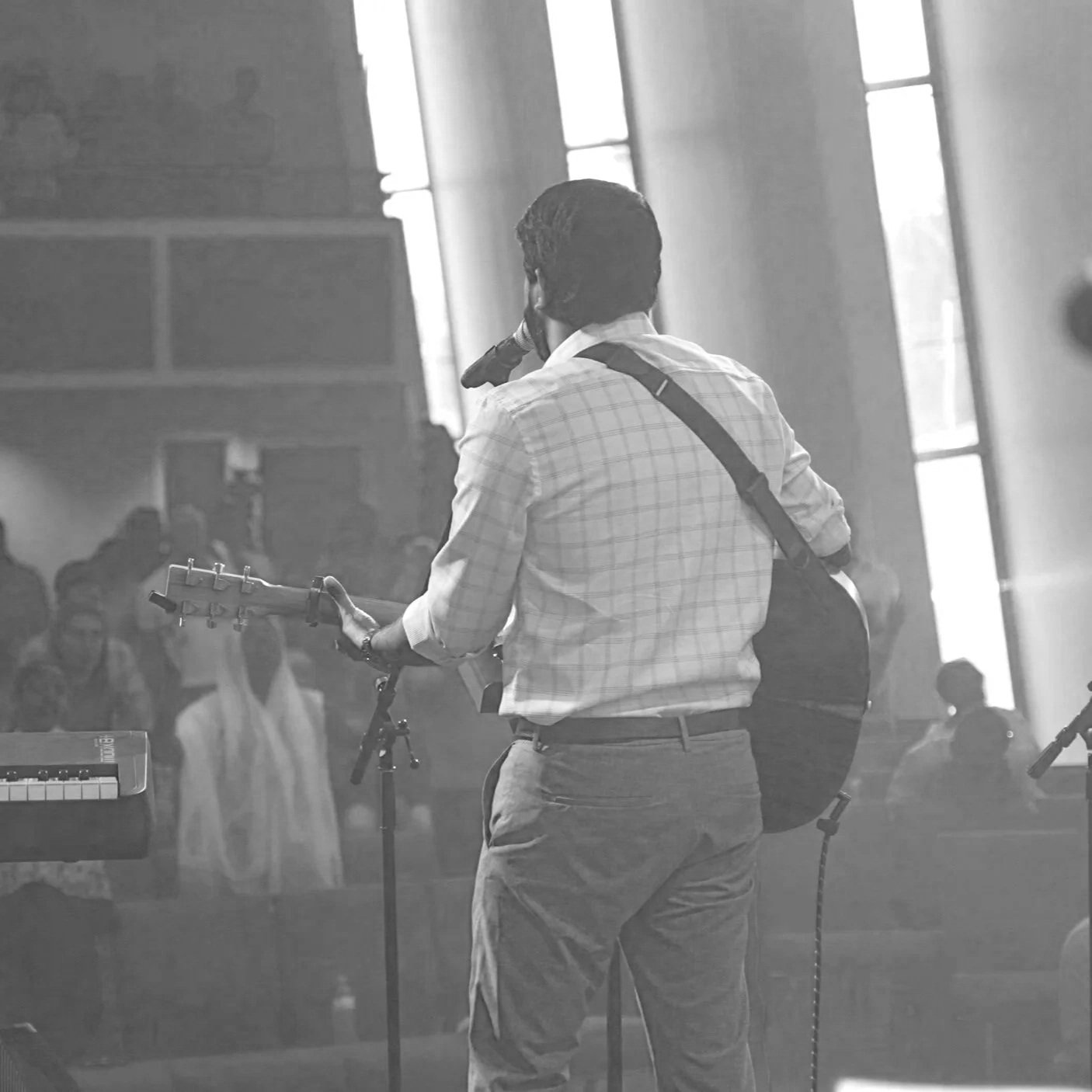A man wearing a checked shirt playing an electric guitar and singing into a microphone during a performance in a church or similar venue with tall windows and soft lighting. There are listeners in the background.