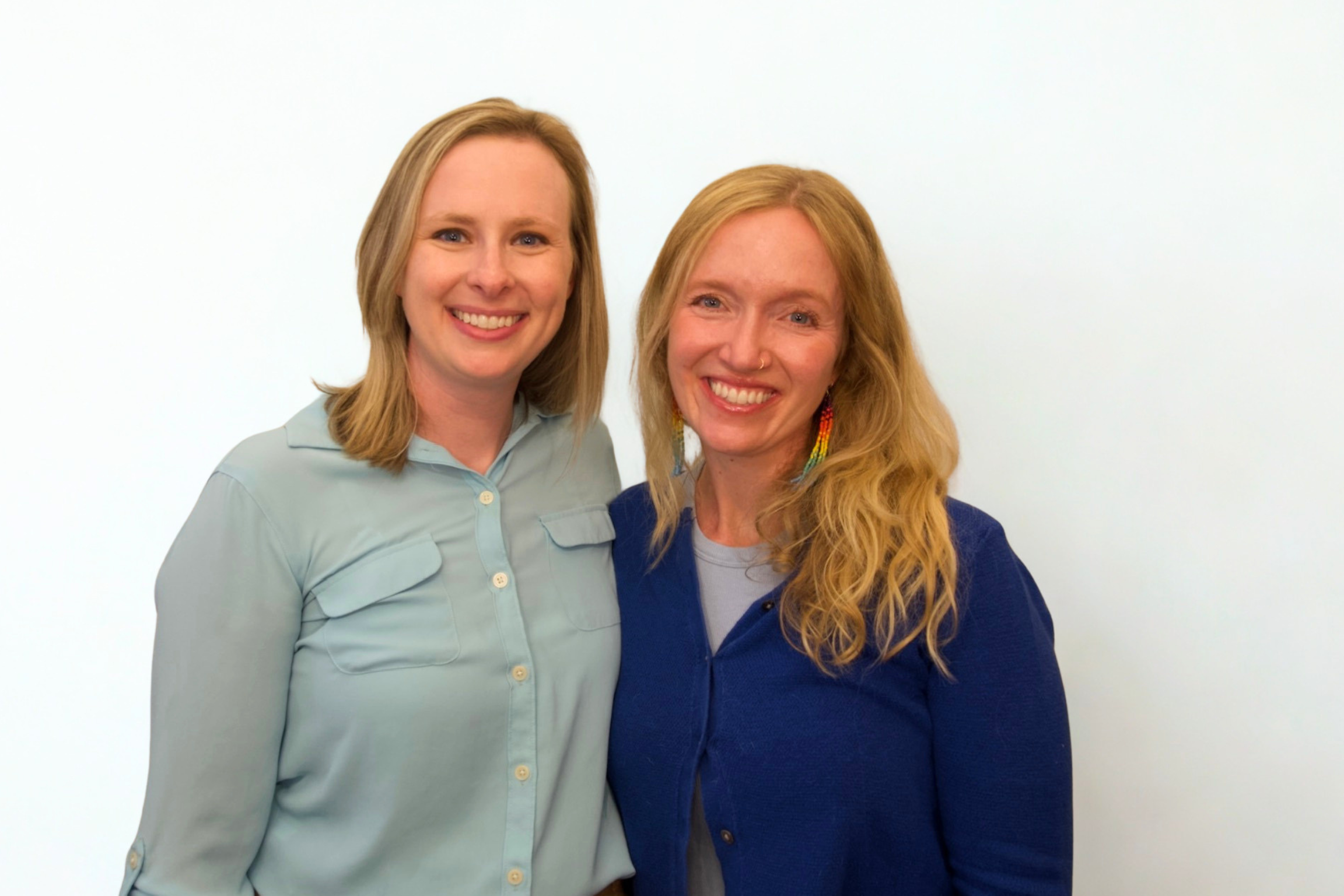 Two women with long, blonde hair standing close together and smiling at the camera against a plain white background.