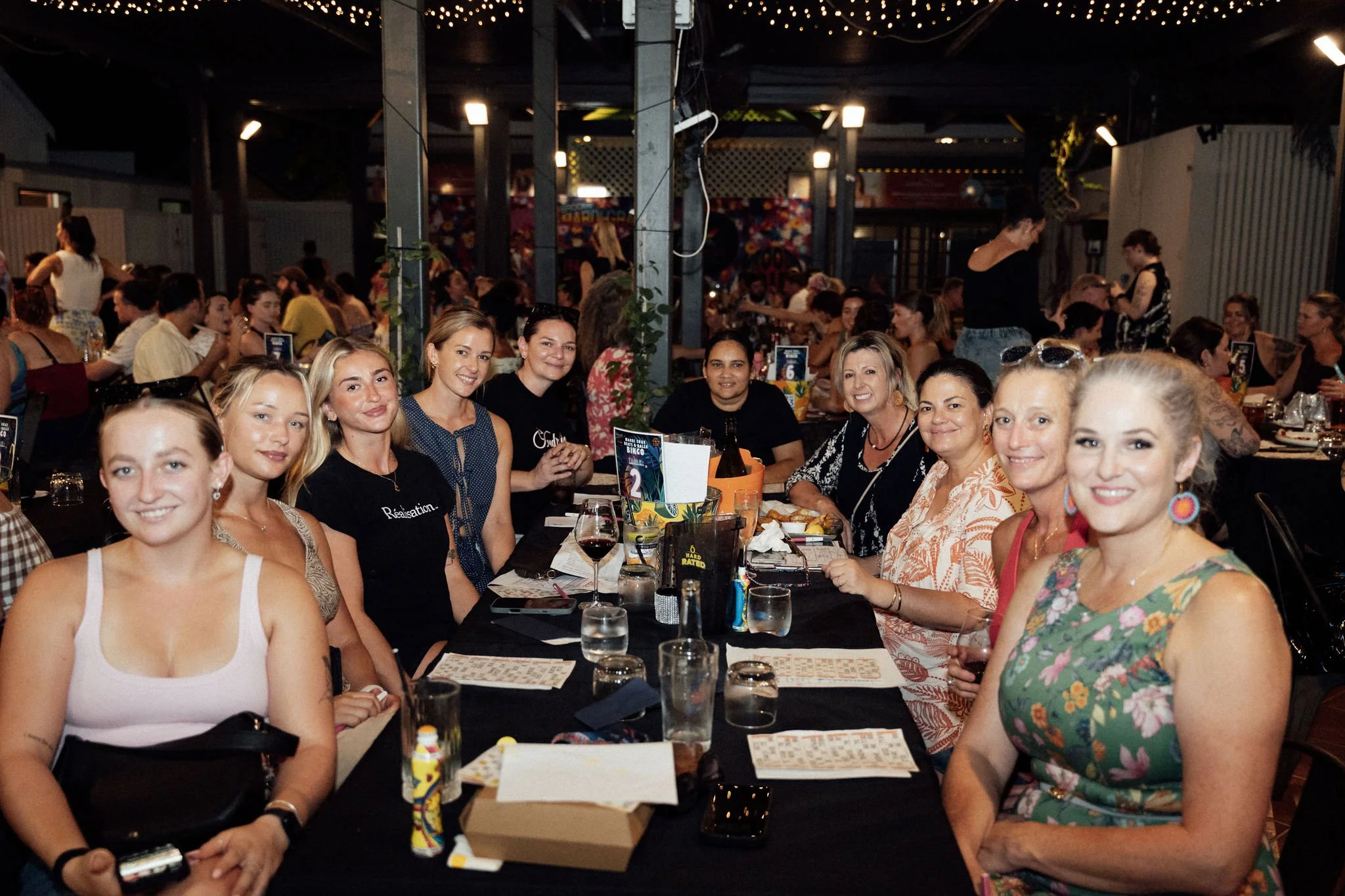 A group of women sitting together at a long table in a dimly lit restaurant or bar, with drinks and menus in front of them. There are more people and high tables in the background, with string lights hanging from the ceiling.
