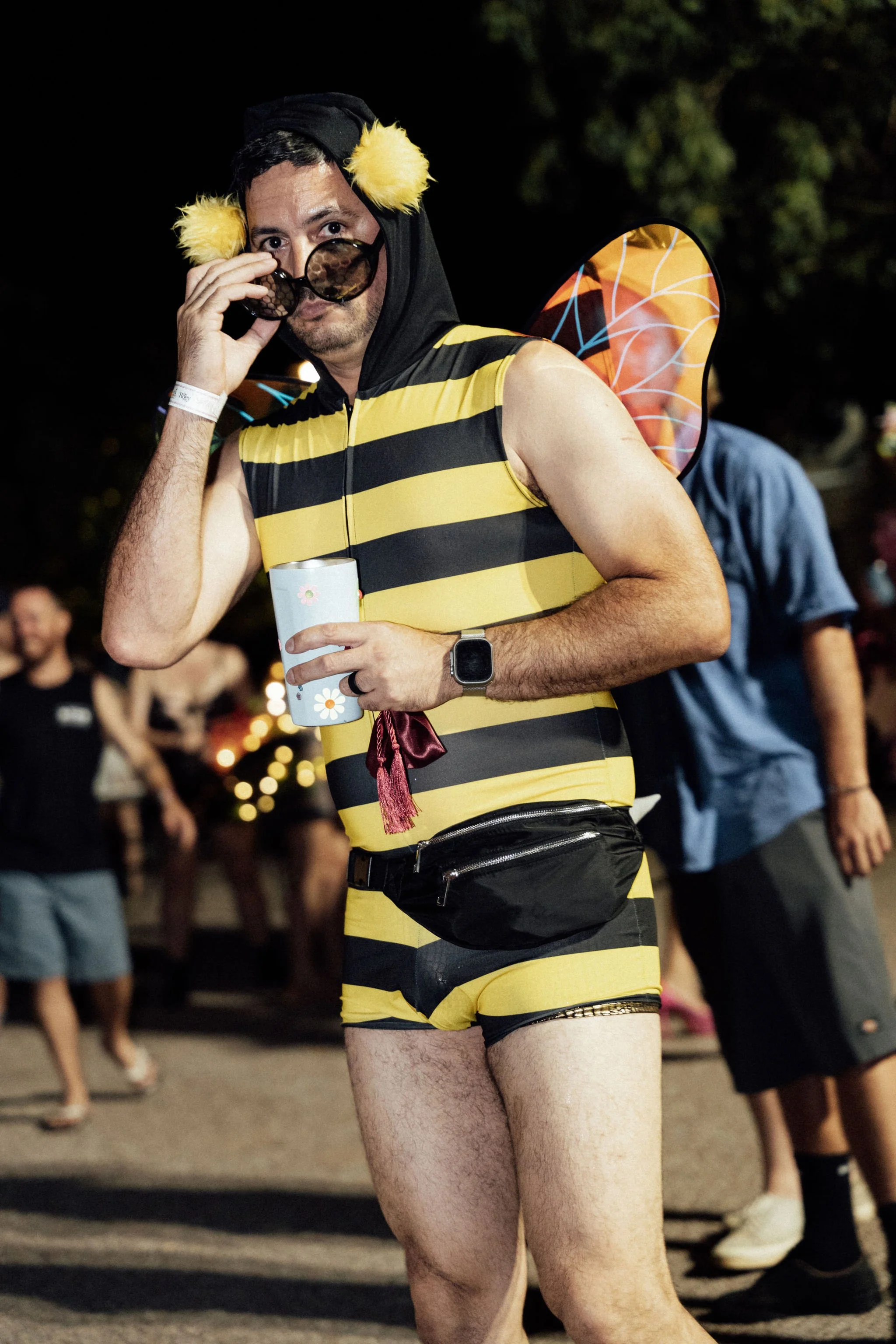 A man wearing a bee costume, including yellow and black striped shorts, a black hood with yellow pom-poms, bee wings, and sunglasses, stands in a crowd at night holding a cup and adjusting his sunglasses.