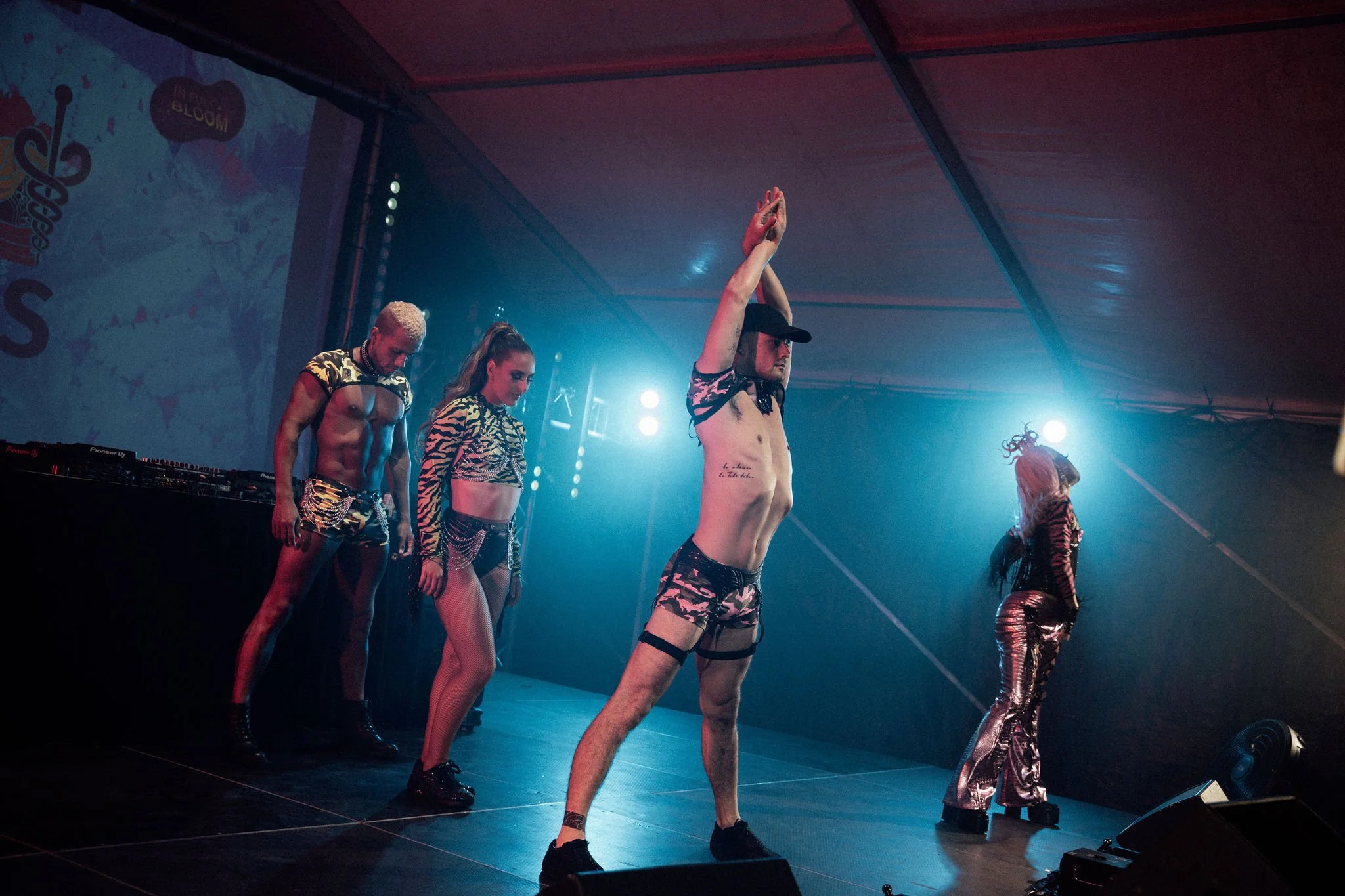 A group of performers on stage wearing leopard print outfits, with some in fishnet stockings, dancing under colorful stage lighting, with a DJ booth on the side.