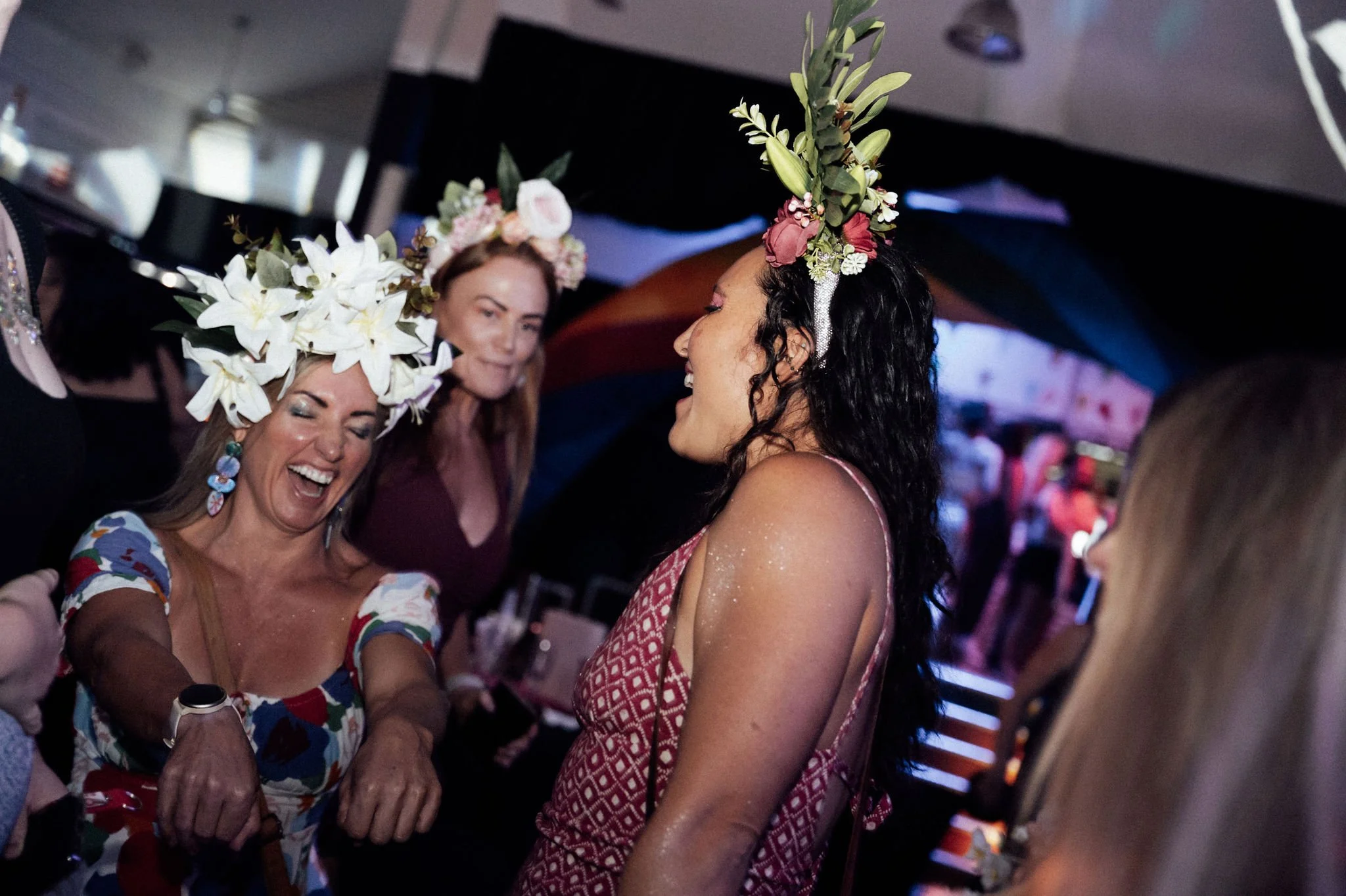 Three women wearing floral headpieces, smiling and celebrating at a social gathering in a dimly lit indoor setting.