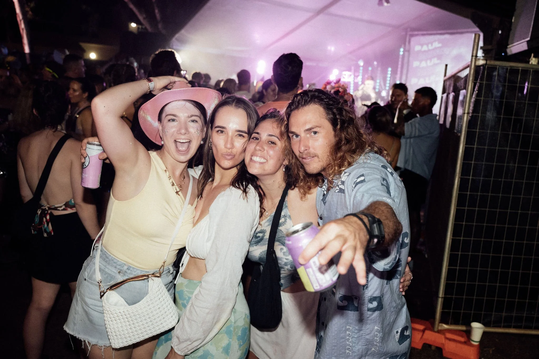 Group of four friends at a music festival, smiling and posing for a selfie, with a large crowd and stage in the background.