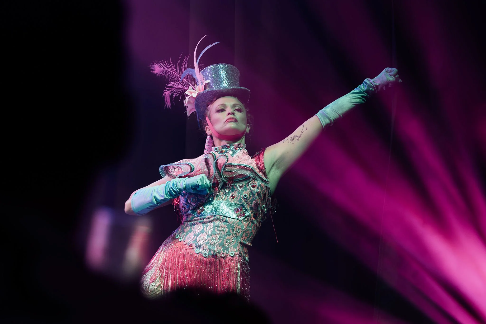 Performer dressed in colorful, glittery costume with a top hat decorated with feathers, striking a pose on stage with purple and pink lighting.