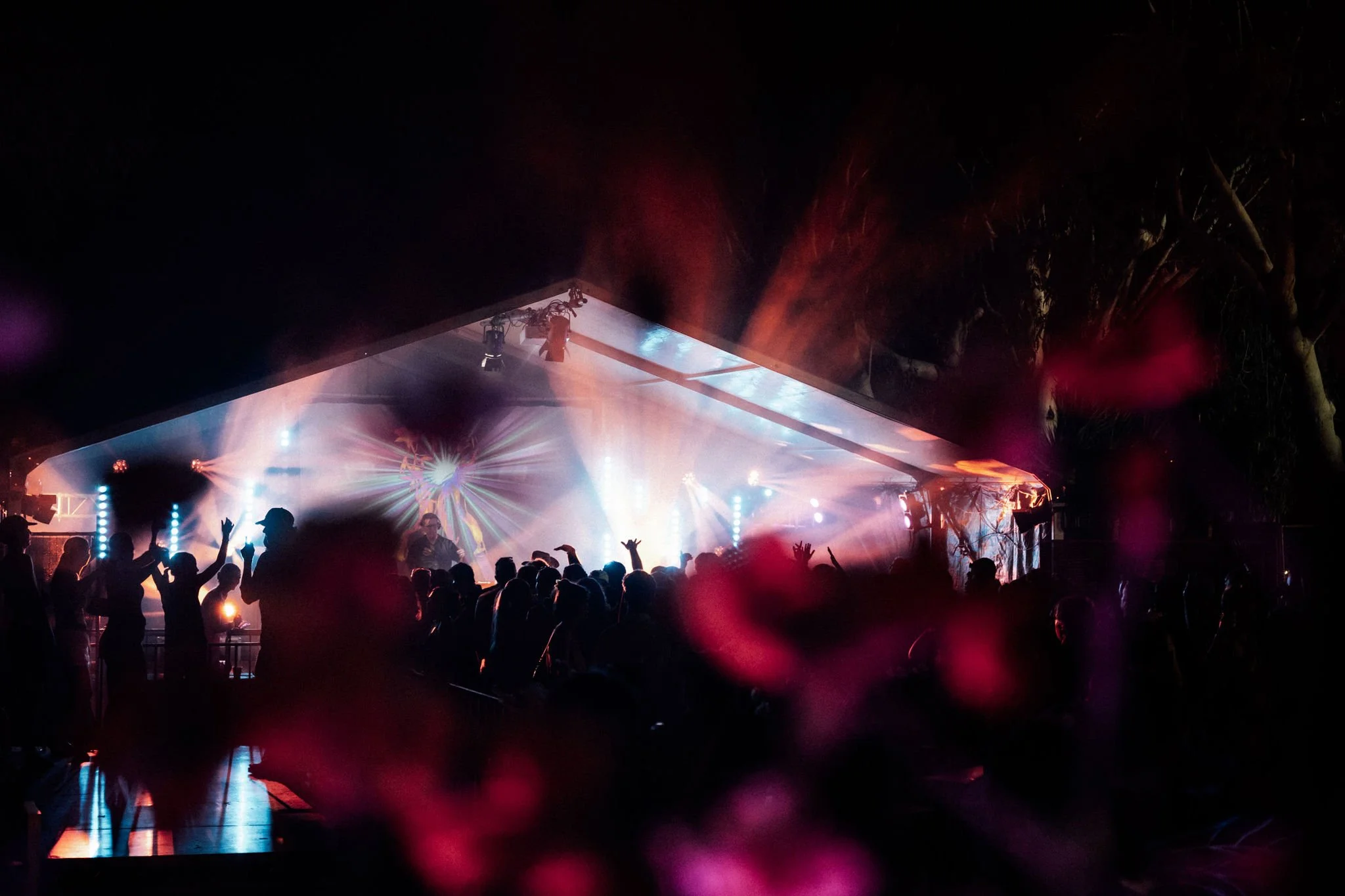People dancing and enjoying a night concert with colorful stage lights, under a canopy, with trees visible in the background.