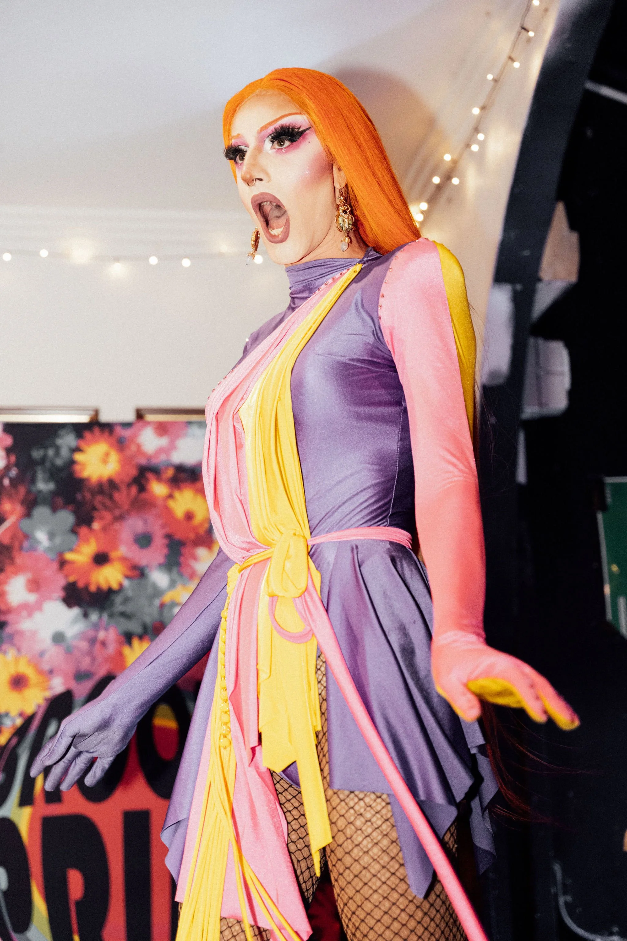 A person with bright orange hair, dramatic makeup, and elaborate earrings is dressed in a colorful, layered outfit with purple, pink, yellow, and fishnet details, appearing to perform or pose.
