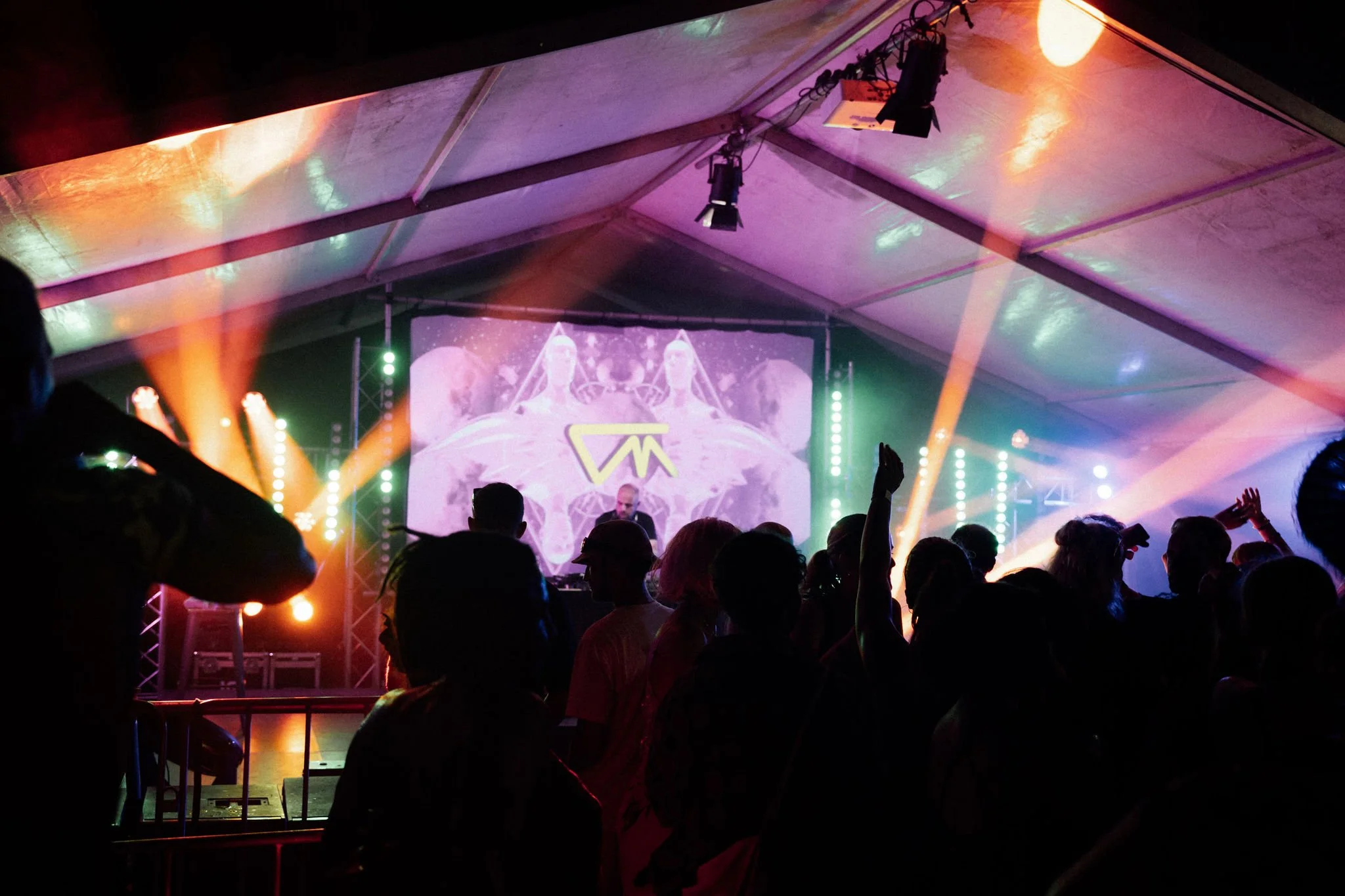 Concert scene with a crowd, colorful stage lighting, and a DJ or performer at the stage in a large tent.