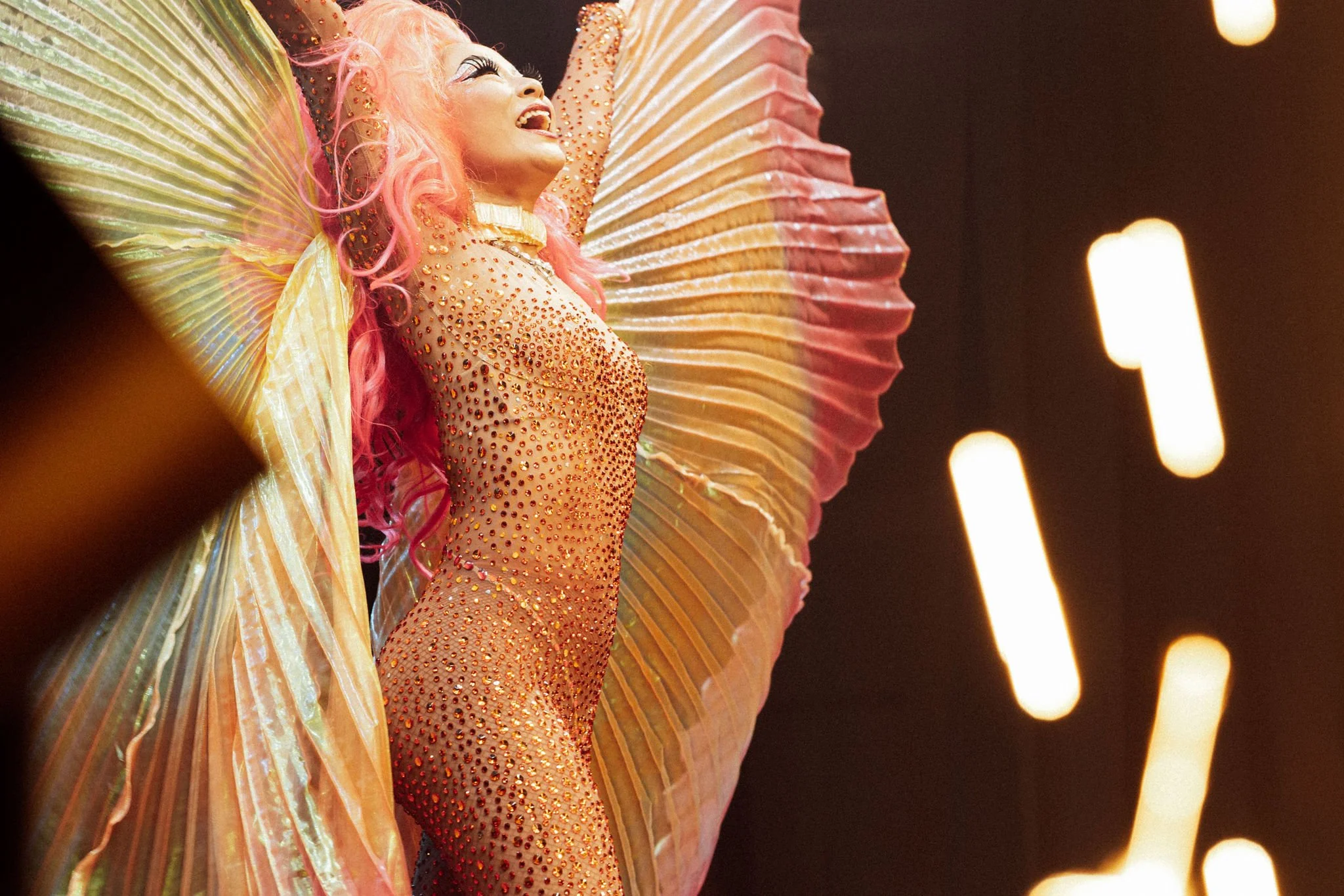 Performer on stage dressed in an elaborate costume with pink hair, rhinestones, and large wing-like accessories, smiling with arms raised.