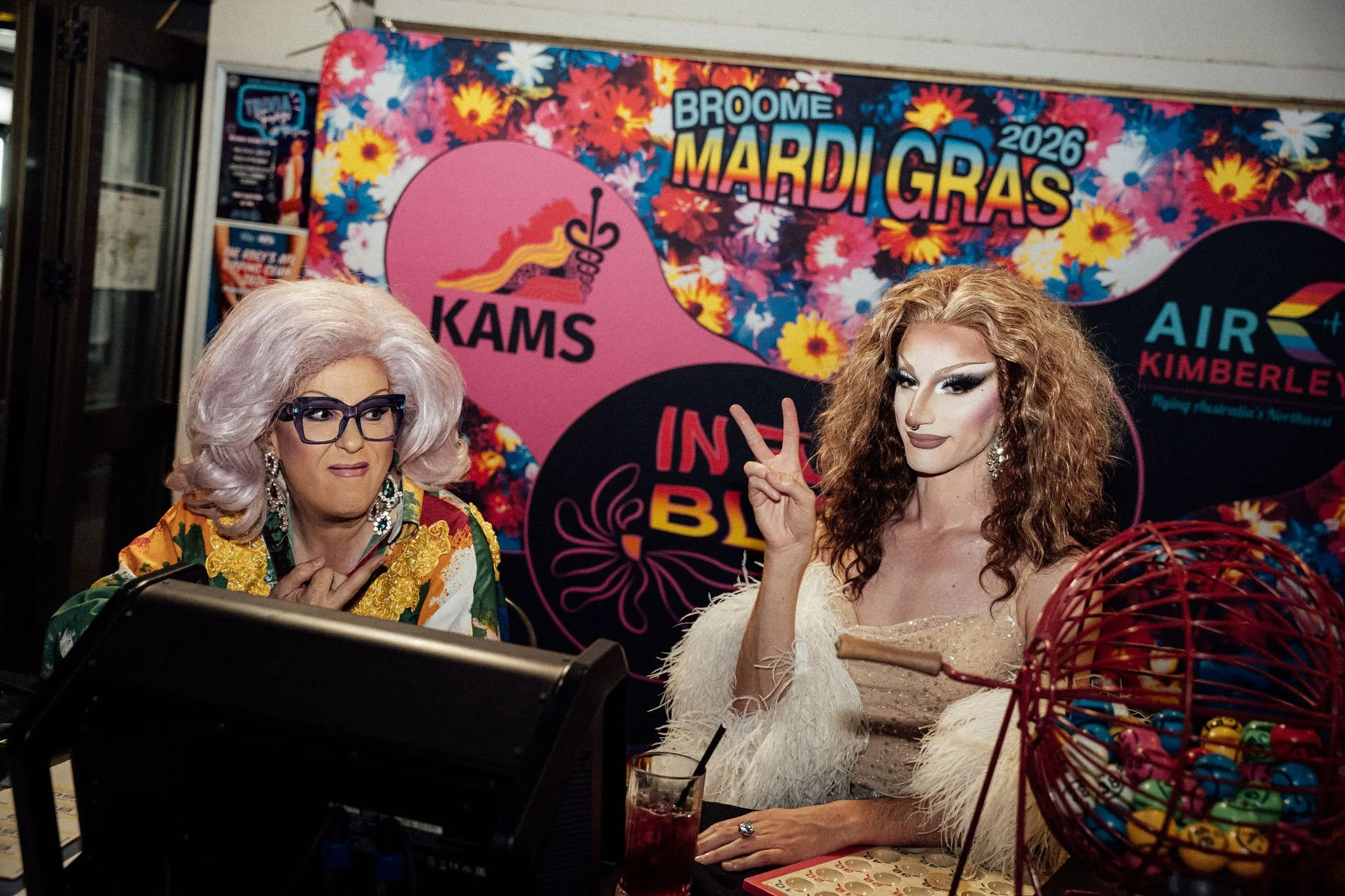 Two drag queens in festive attire seated at a table with colorful baskets of candy, with a vibrant floral and event banner in the background celebrating Mardi Gras 2026.