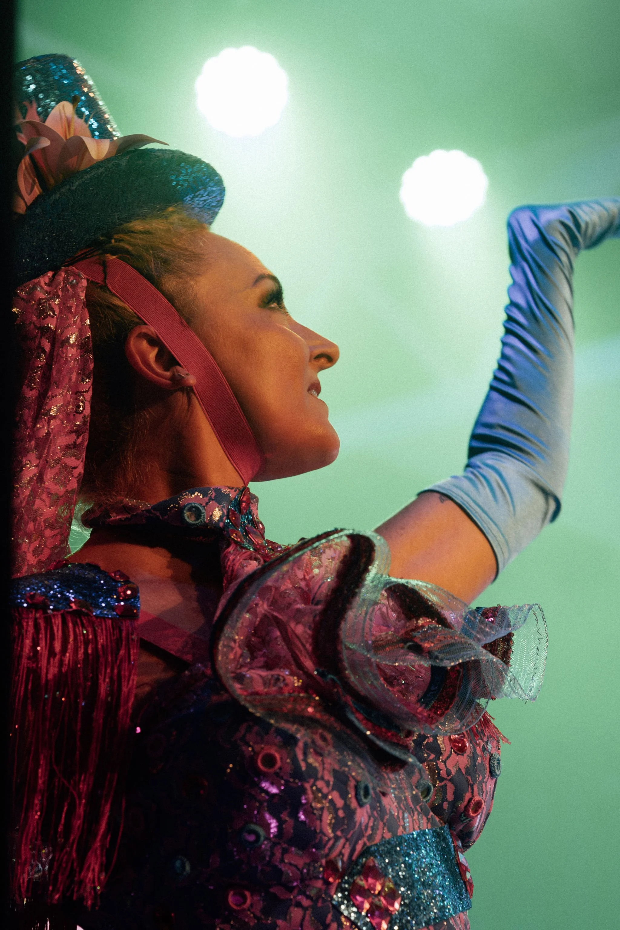 A performer with long blond hair wearing a colorful, sequined and embroidered costume with ruffles and fringes, raising one arm, with a green background and bright lights.