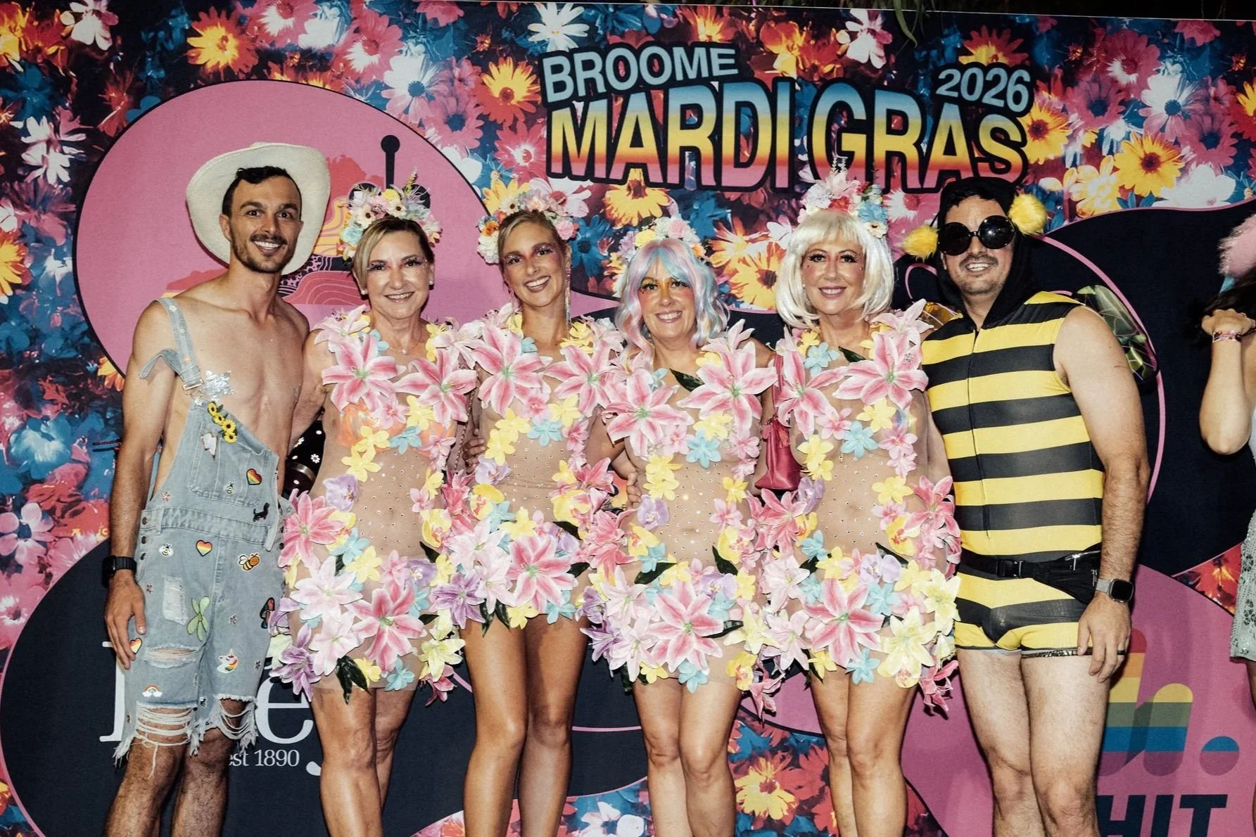 Group of six people dressed in colorful, festive costumes standing in front of a floral backdrop with the text 'Broome Mardi Gras 2026' and a pink circle with design elements.