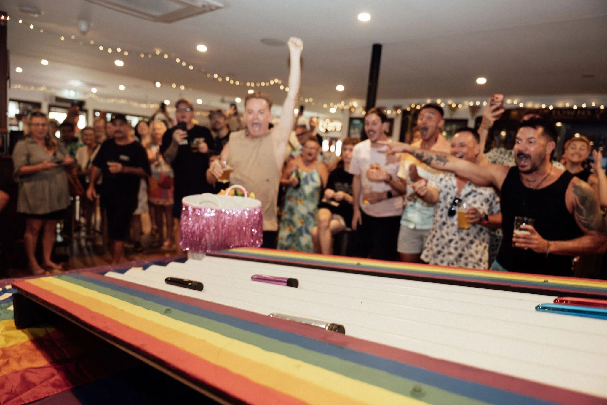 Group of people celebrating around a table with a rainbow-colored cake and cake topper, one person is raising their arm in excitement, some people are holding drinks, in a decorated indoor setting with fairy lights.