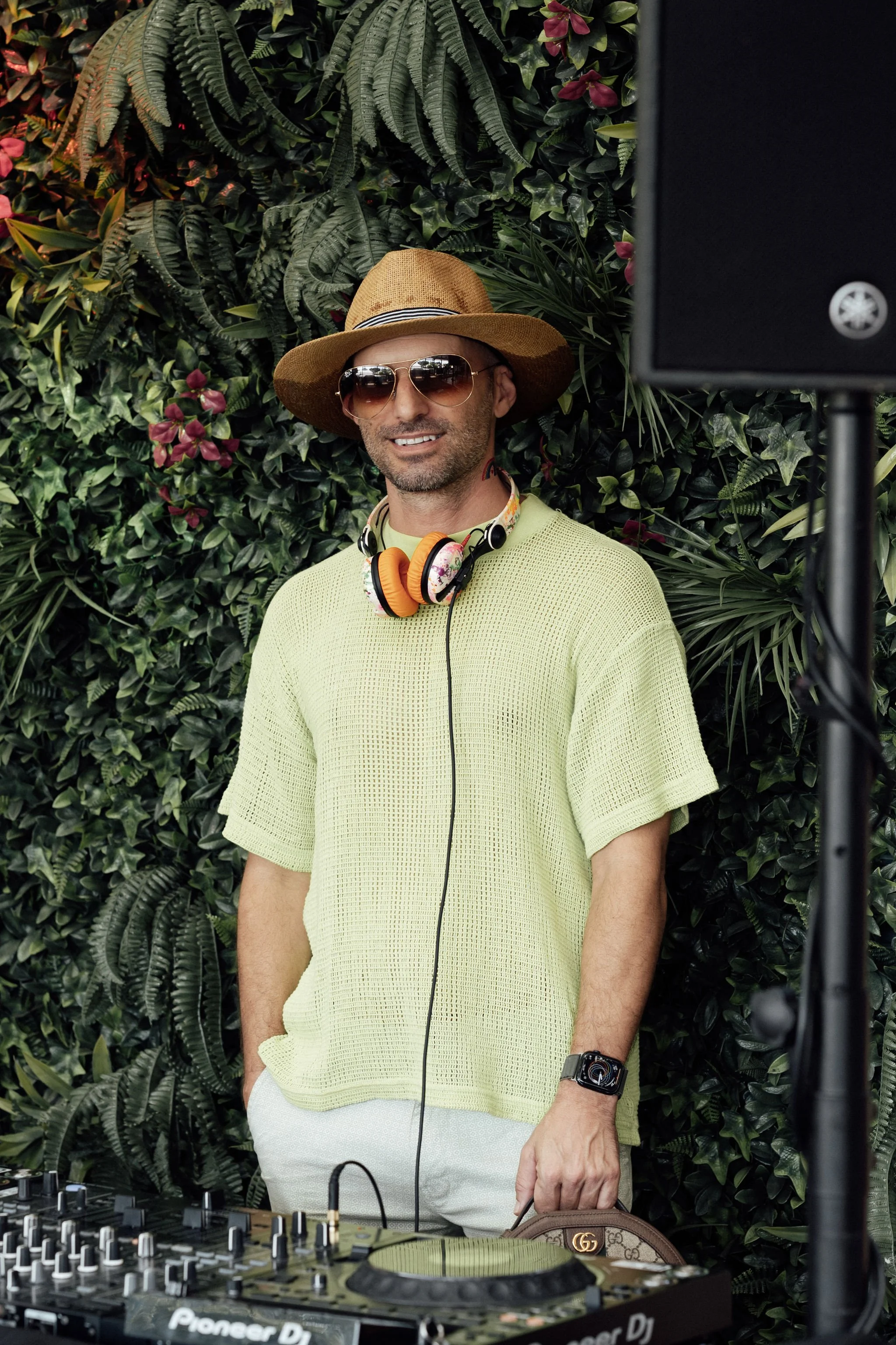 A man is smiling while standing behind DJ equipment, wearing sunglasses, a straw hat, a yellow mesh shirt, and white shorts, with a lush green and pink flower backdrop.