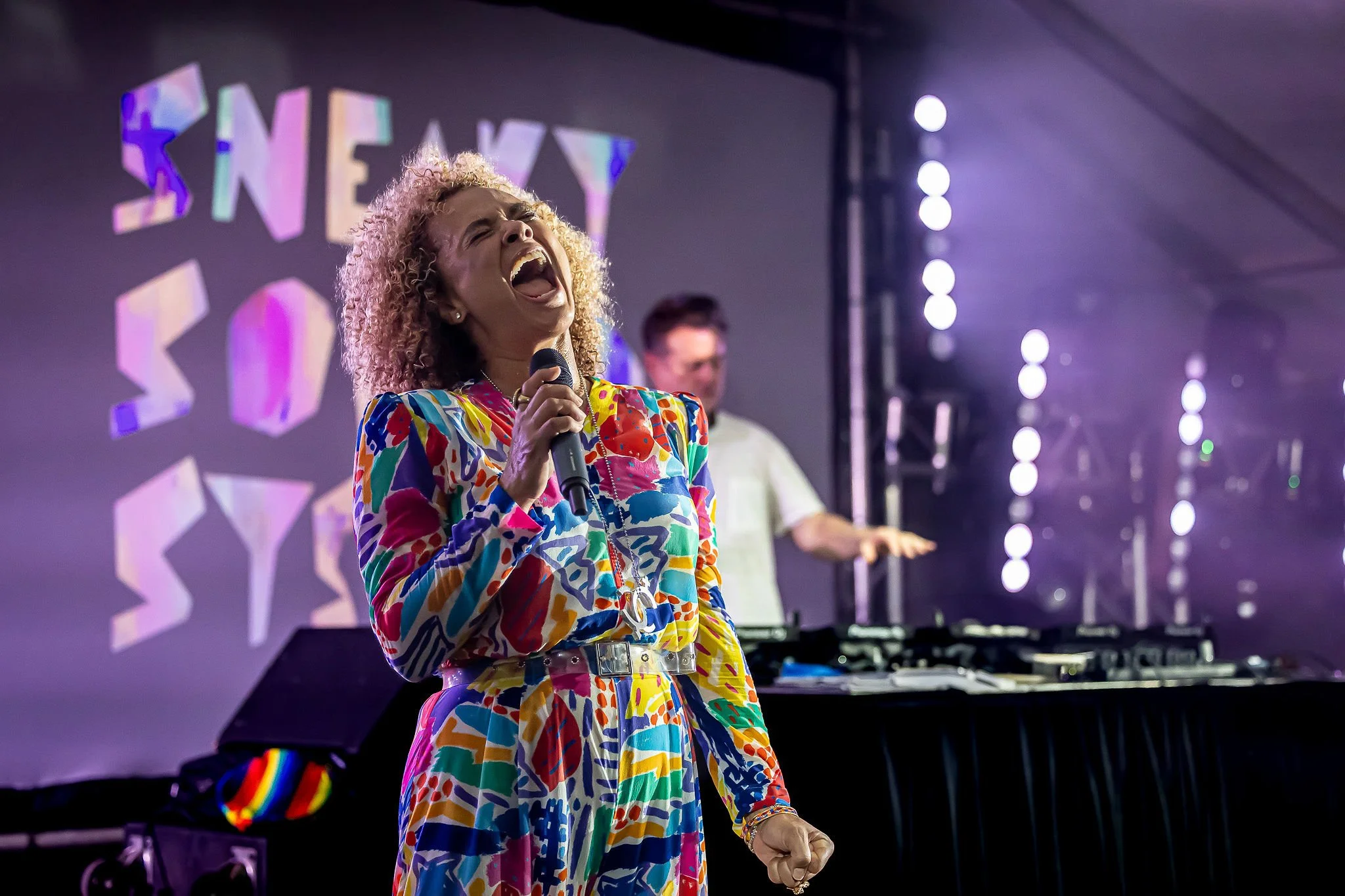 A woman with curly blonde hair passionately singing into a microphone during a stage performance, wearing a colorful patterned dress, with a background that includes a person at a keyboard and stage lights.