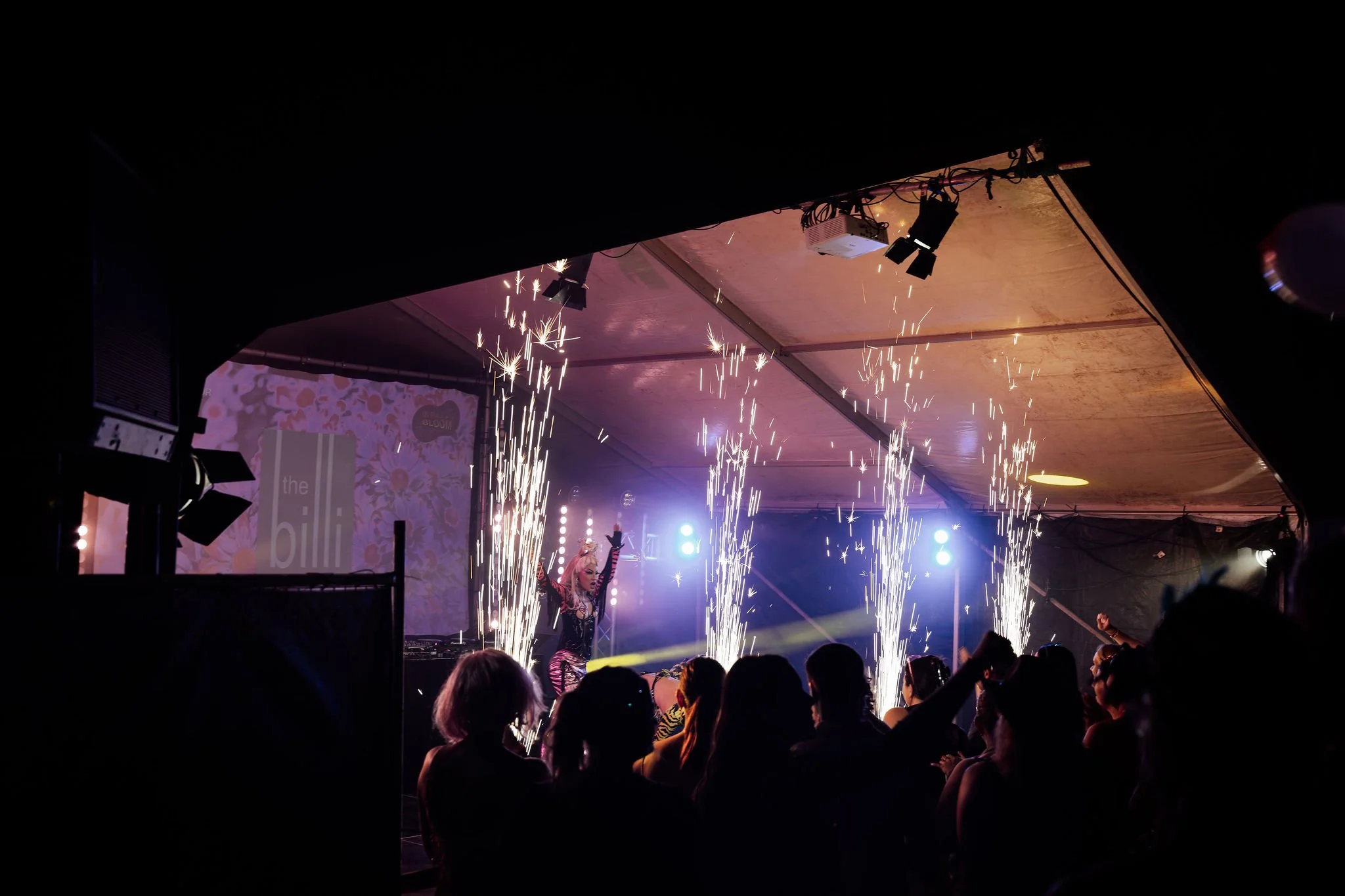 A performer on stage wearing a costume with long gloves and a headpiece, with sparks and pyrotechnics creating a fiery display while an audience watches in a darkened setting.