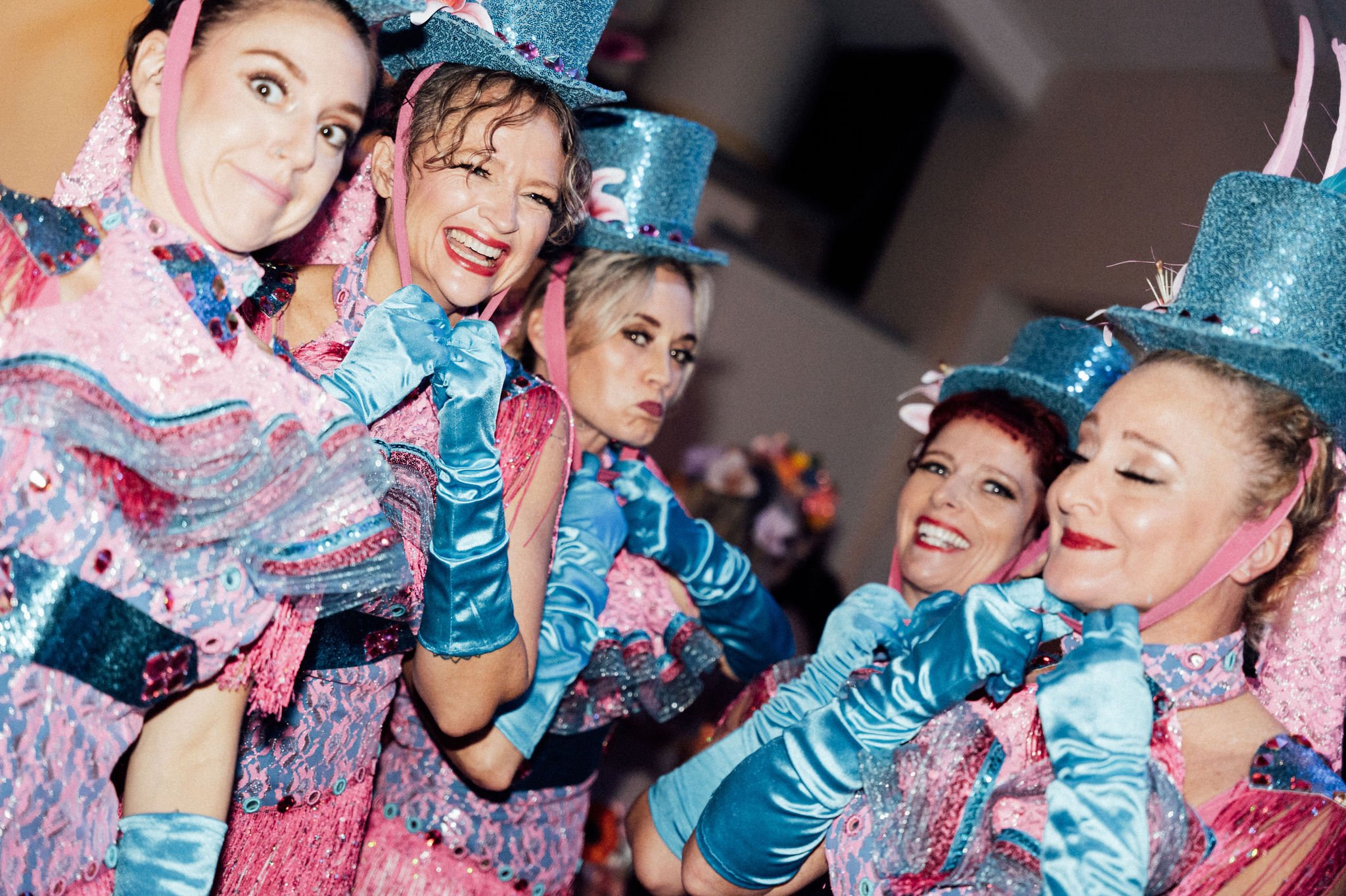 Group of women dressed in colorful, sparkly costumes with blue hats, smiling and interacting during a celebration or performance.