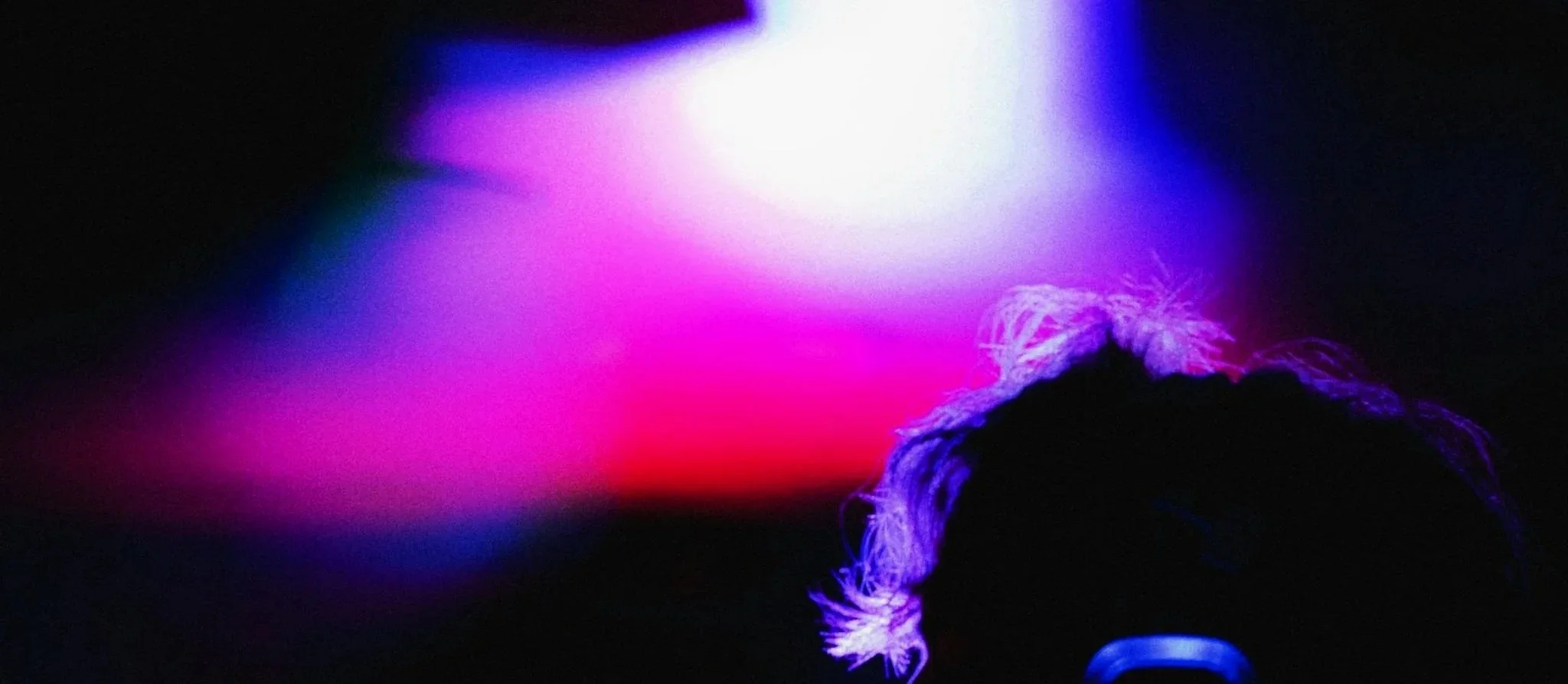 A dark scene with vibrant pink, purple, blue, and white lights creating an abstract background. The silhouette of a person with curly hair is visible in the foreground, facing away from the camera.