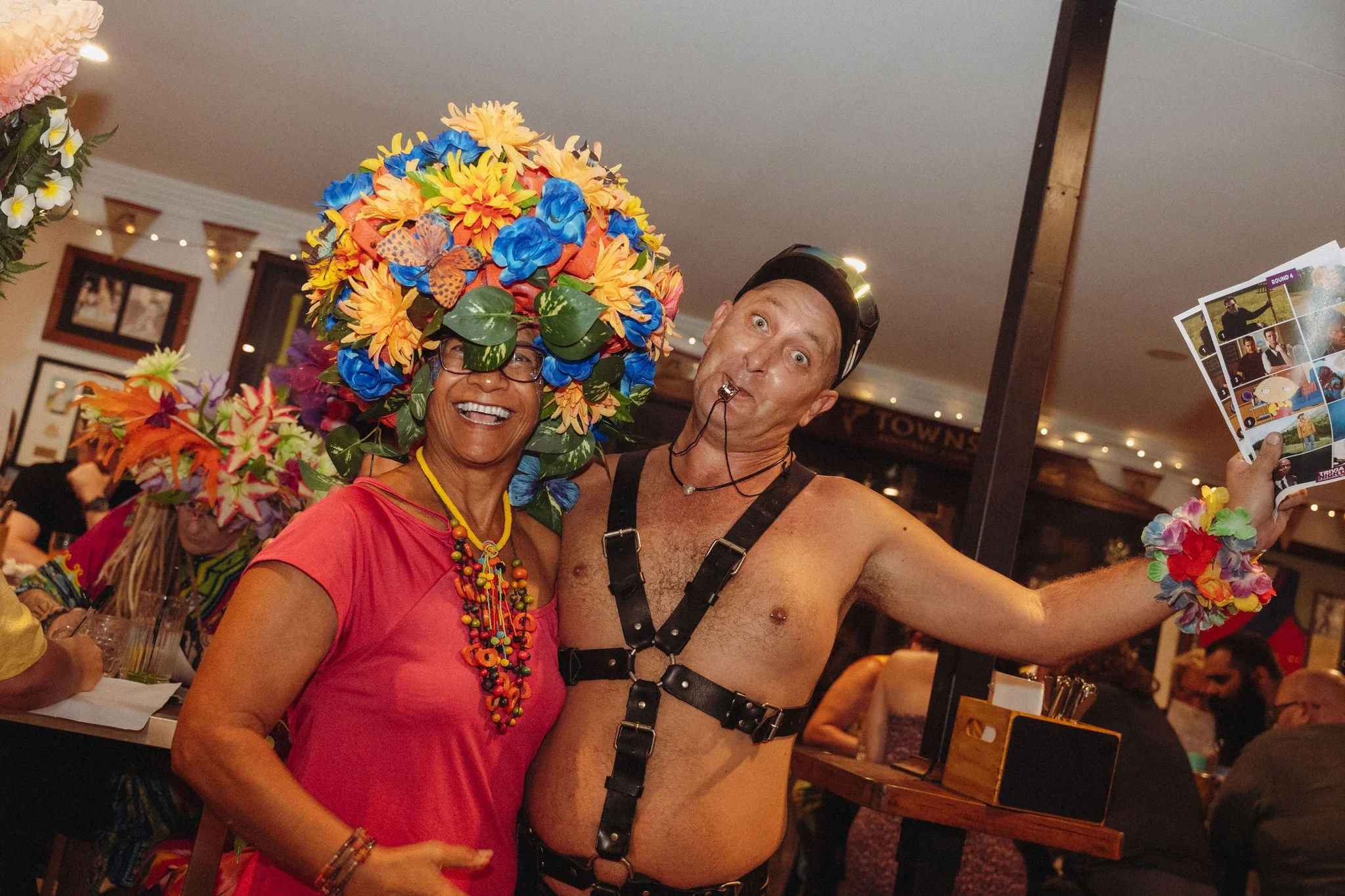 A woman in a bright pink dress and colorful necklace, wearing a large floral headpiece, smiling beside a shirtless man dressed as a bondage character, both taking a selfie at a festive indoor event.