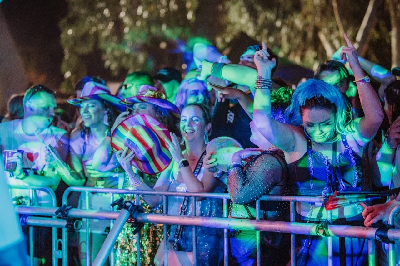 Crowd of people dancing at a nighttime outdoor event, illuminated by colorful lights, with some wearing hats and vibrant costumes.