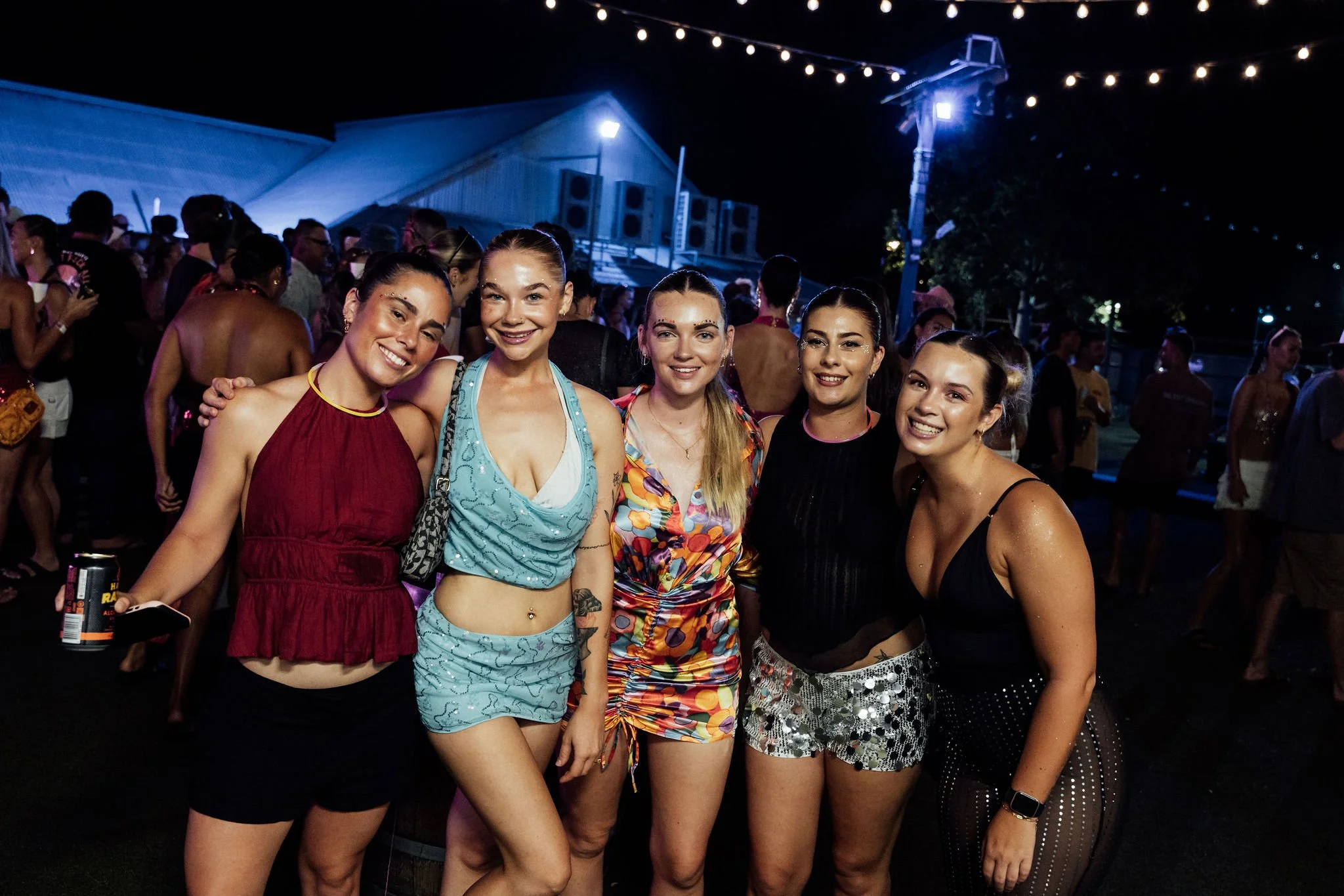 Four young women standing together at a night outdoor event, smiling at the camera, with a crowd of people and string lights in the background.