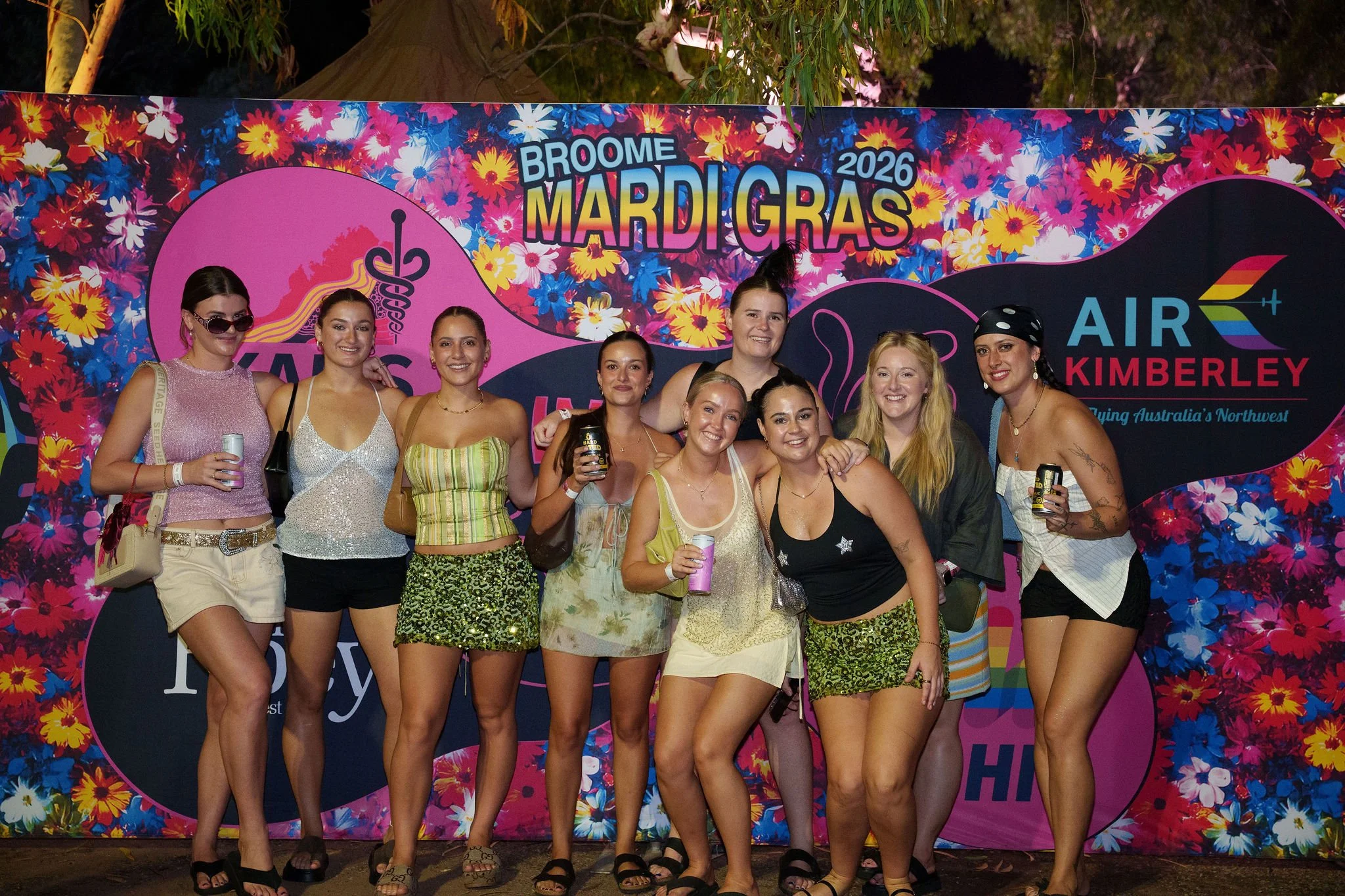 A group of nine women posing at a Mardi Gras event in 2026, standing in front of a colorful floral backdrop with Mardi Gras themed text and symbols.