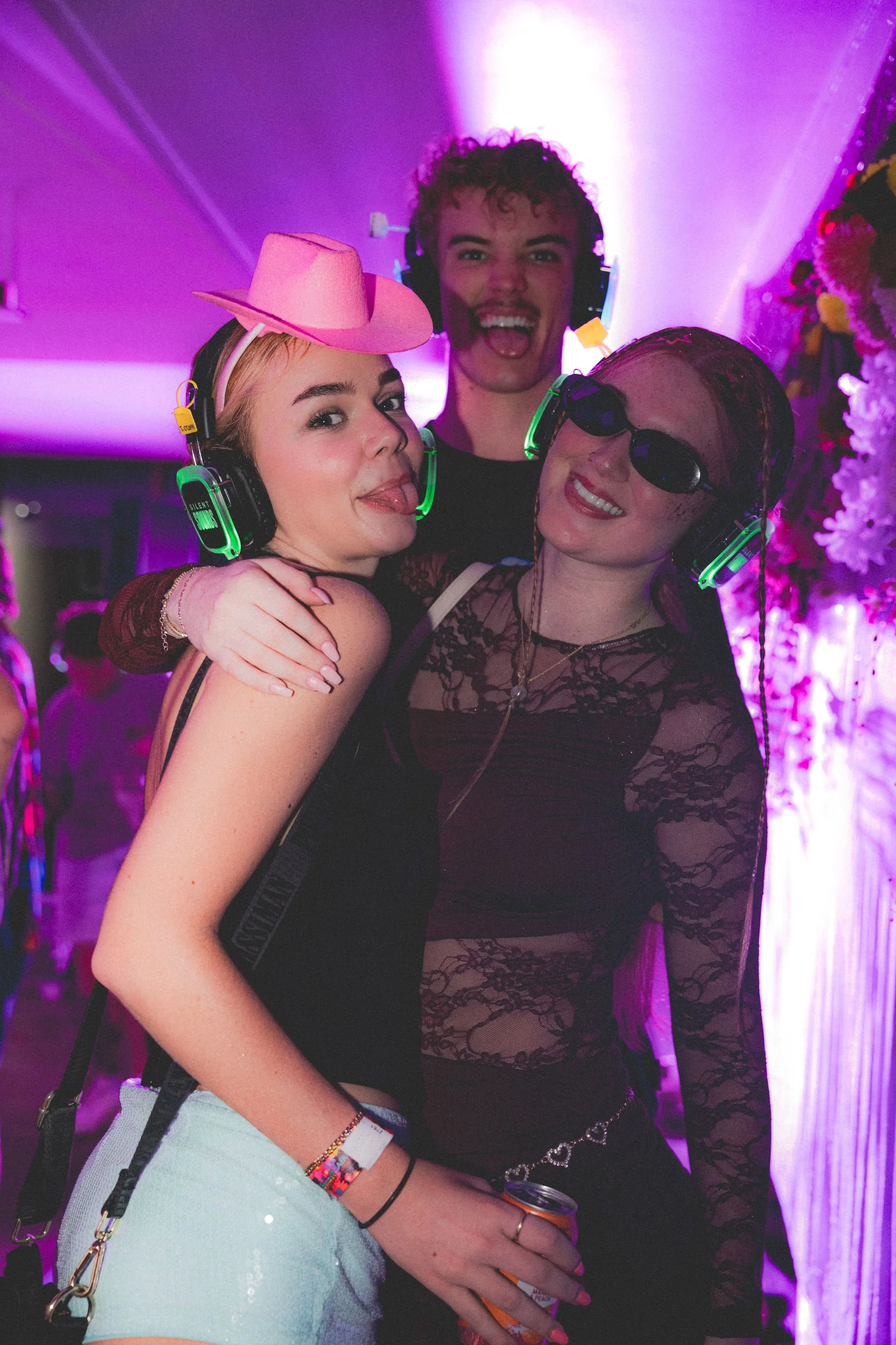 Three young women at a party wearing headphones with neon lights, one with a pink cowboy hat and sticking out her tongue, another with sunglasses and a nose ring, and the third with a neck chain, dressed in black lace, smiling and having fun.