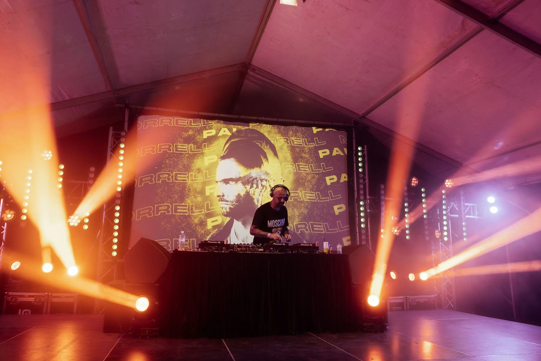A DJ wearing a Moschino t-shirt performing on stage with a large LED screen displaying the face of a character from the movie Predator in the background. The stage is illuminated with bright orange and purple lights.
