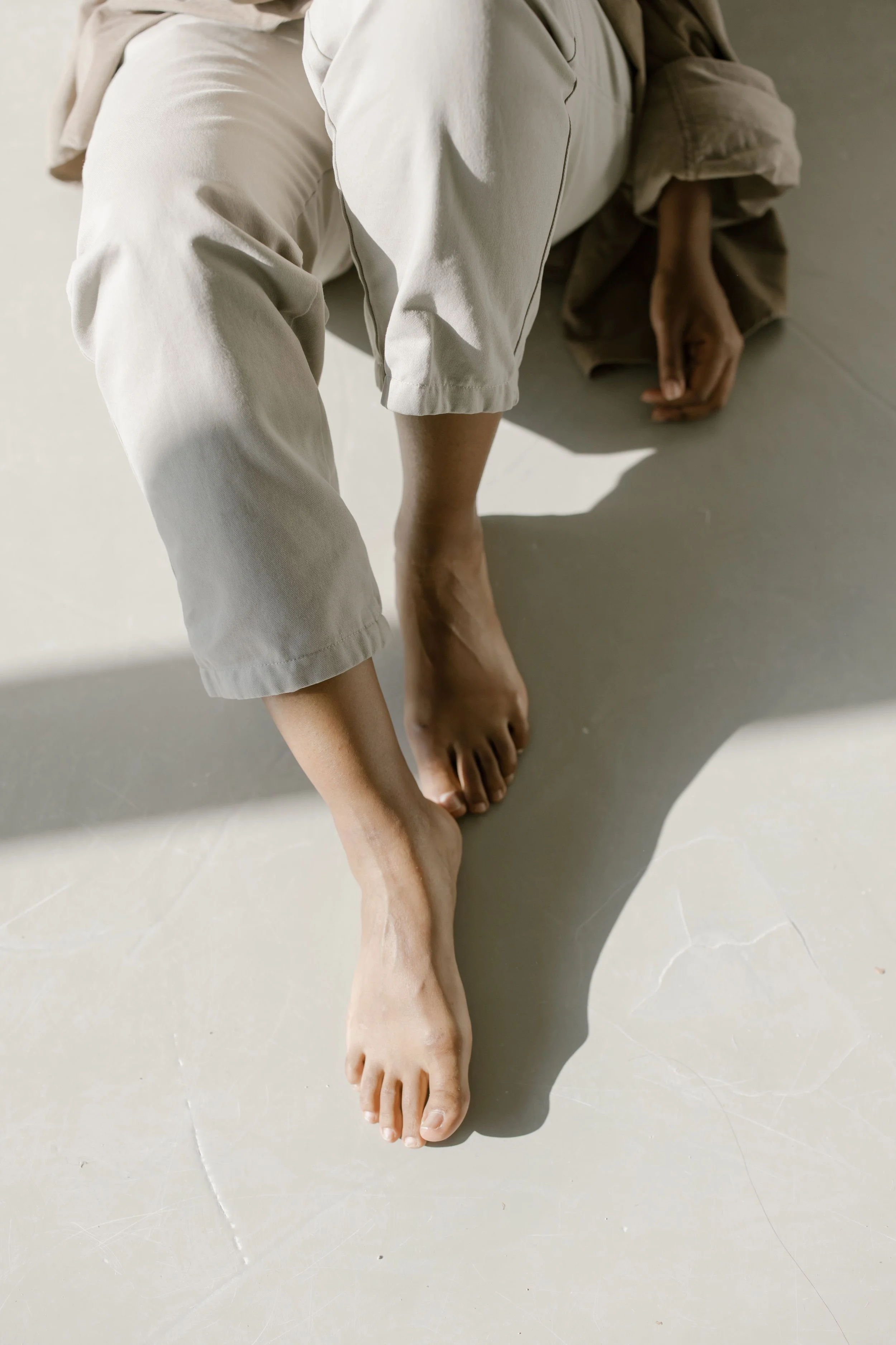 Person wearing beige pants and shirt, sitting on a neutral-colored floor, with one arm extended and hand resting on the floor.