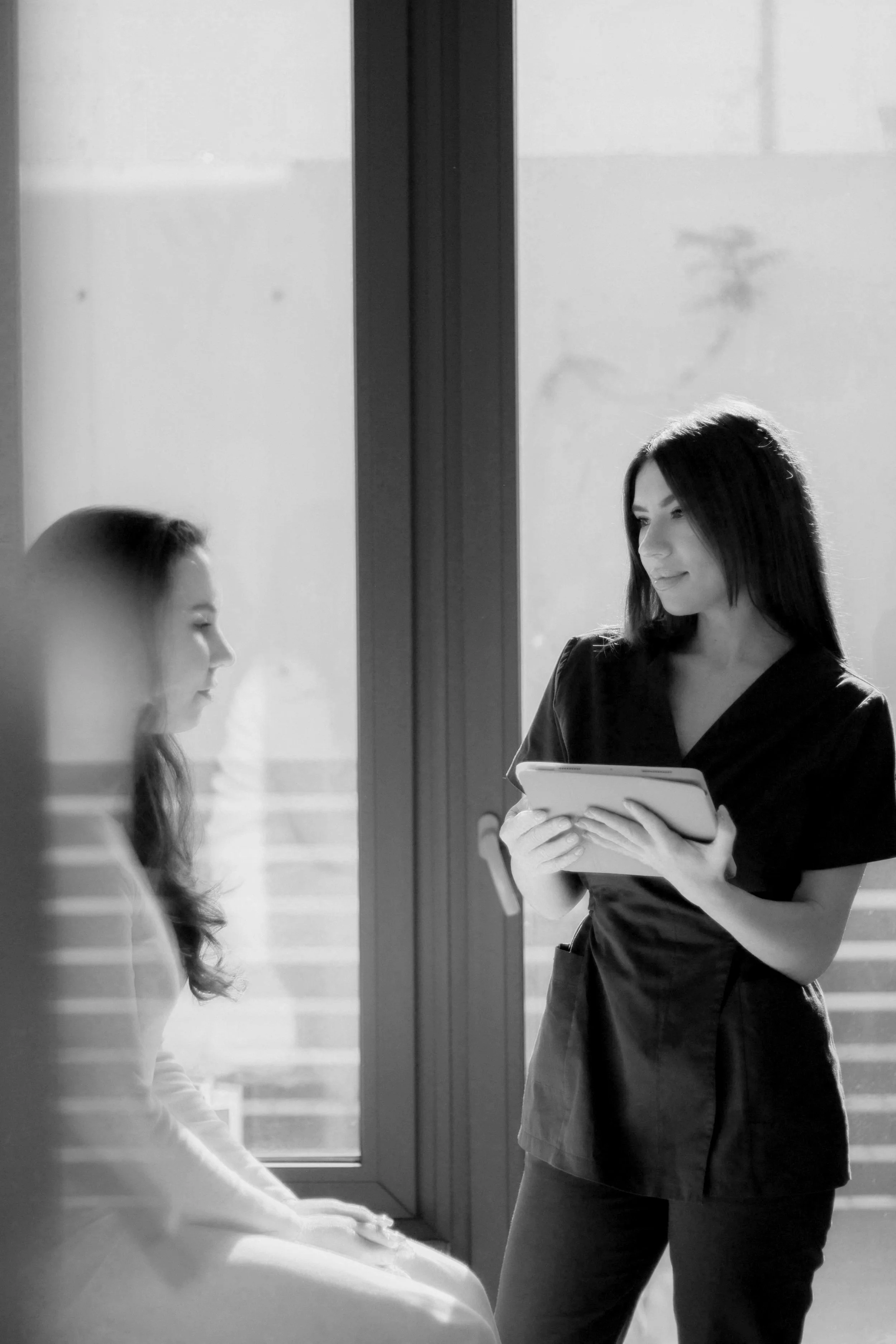 A woman holding a tablet is talking to a seated woman near a large window; the scene is in black and white.