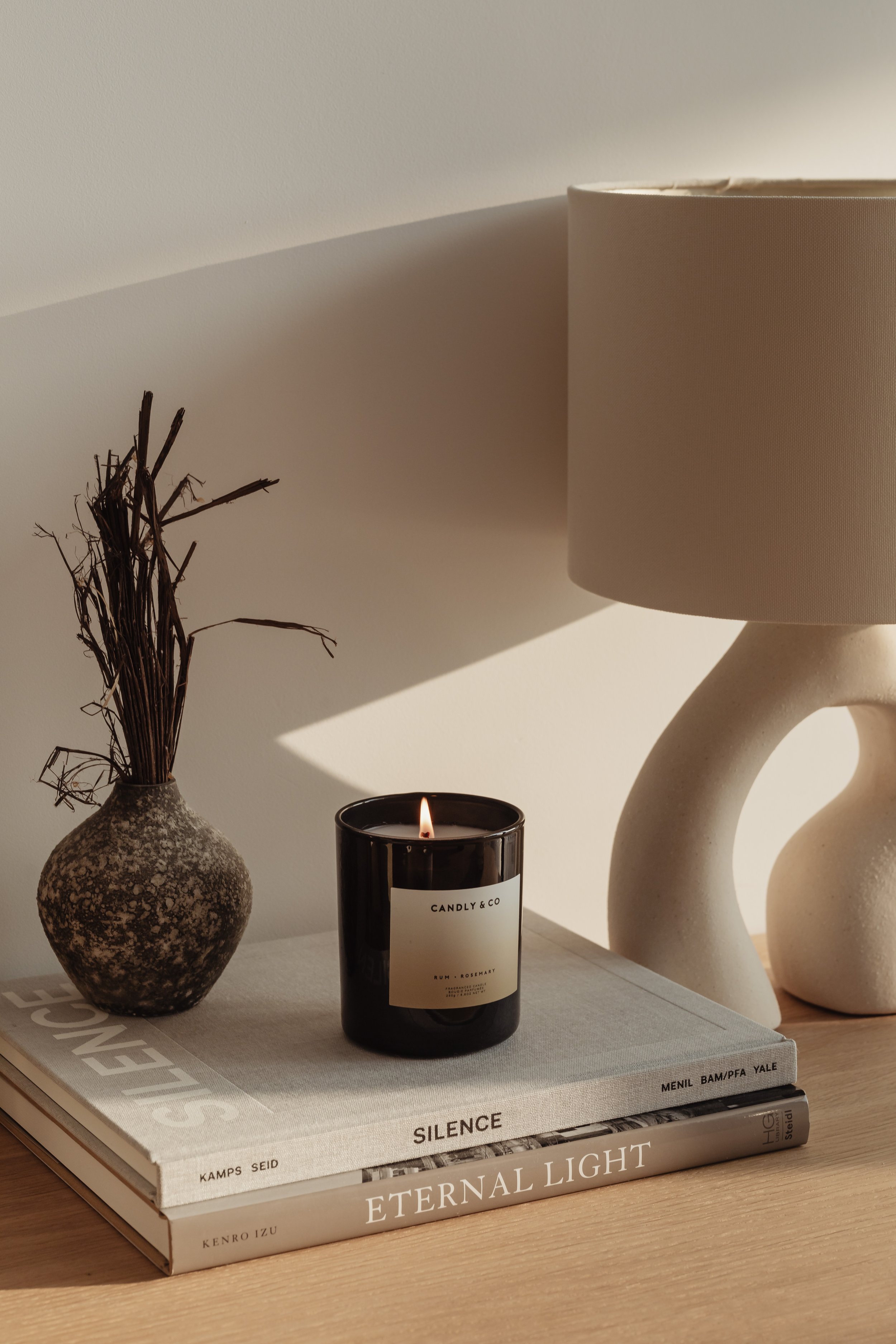 A beige table lamp, a dried branch arrangement in a textured ceramic vase, a black candle with a white label, and stacked books on a wooden surface.
