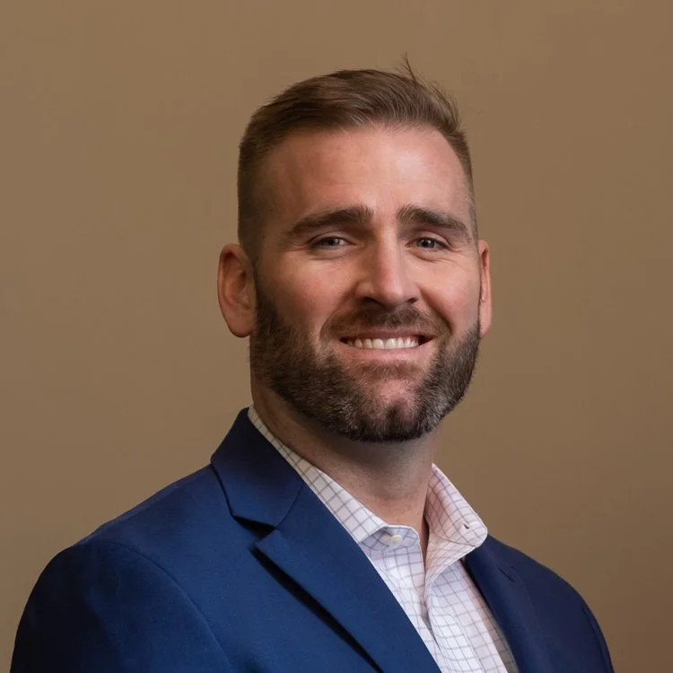 Portrait of a smiling man with a beard, wearing a navy blazer and a light checkered shirt, against a plain background.