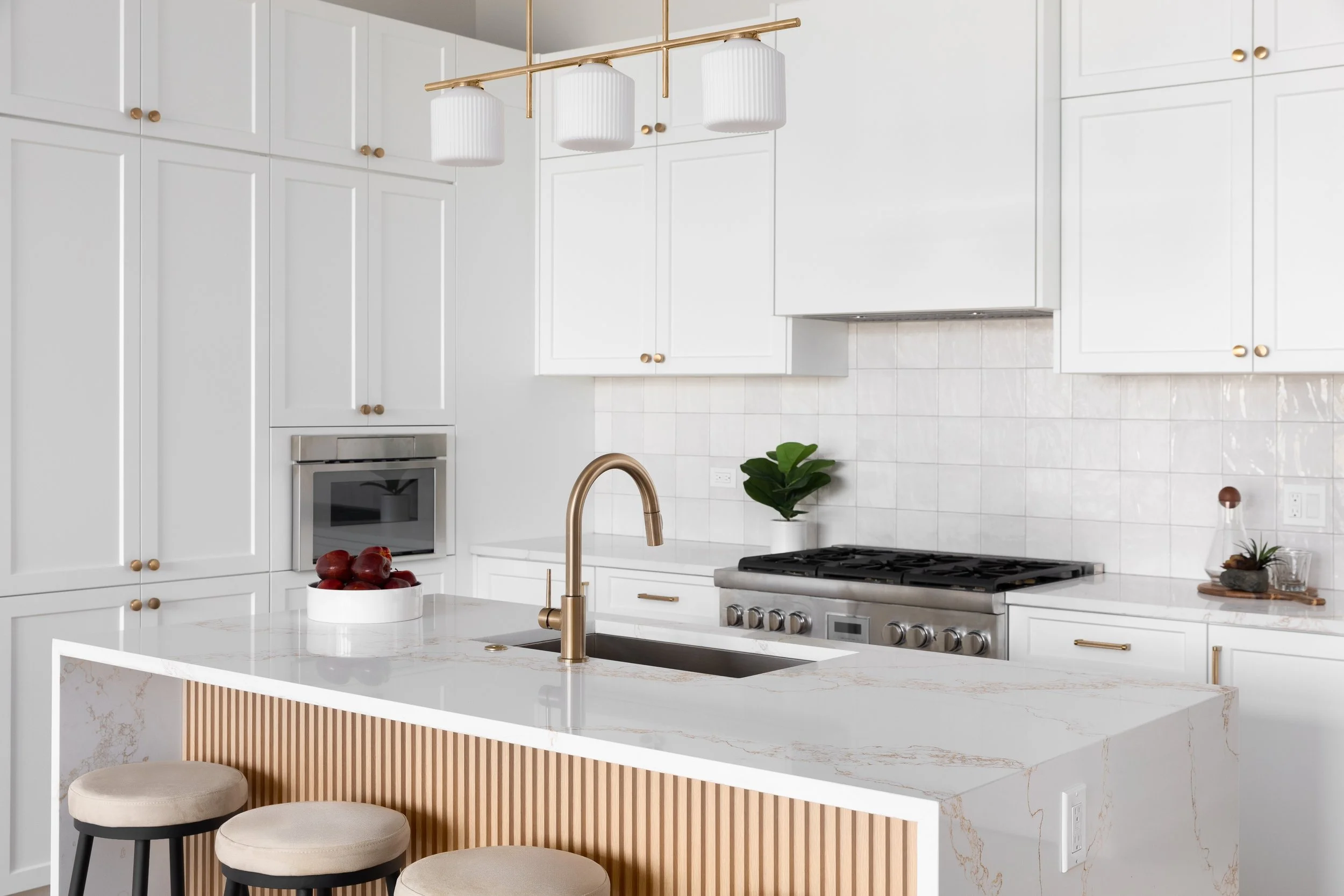 White modern kitchen with gold accents, featuring white cabinets, a white marble countertop island with a table and three beige stools, a built-in oven, a plant, and decorative items.