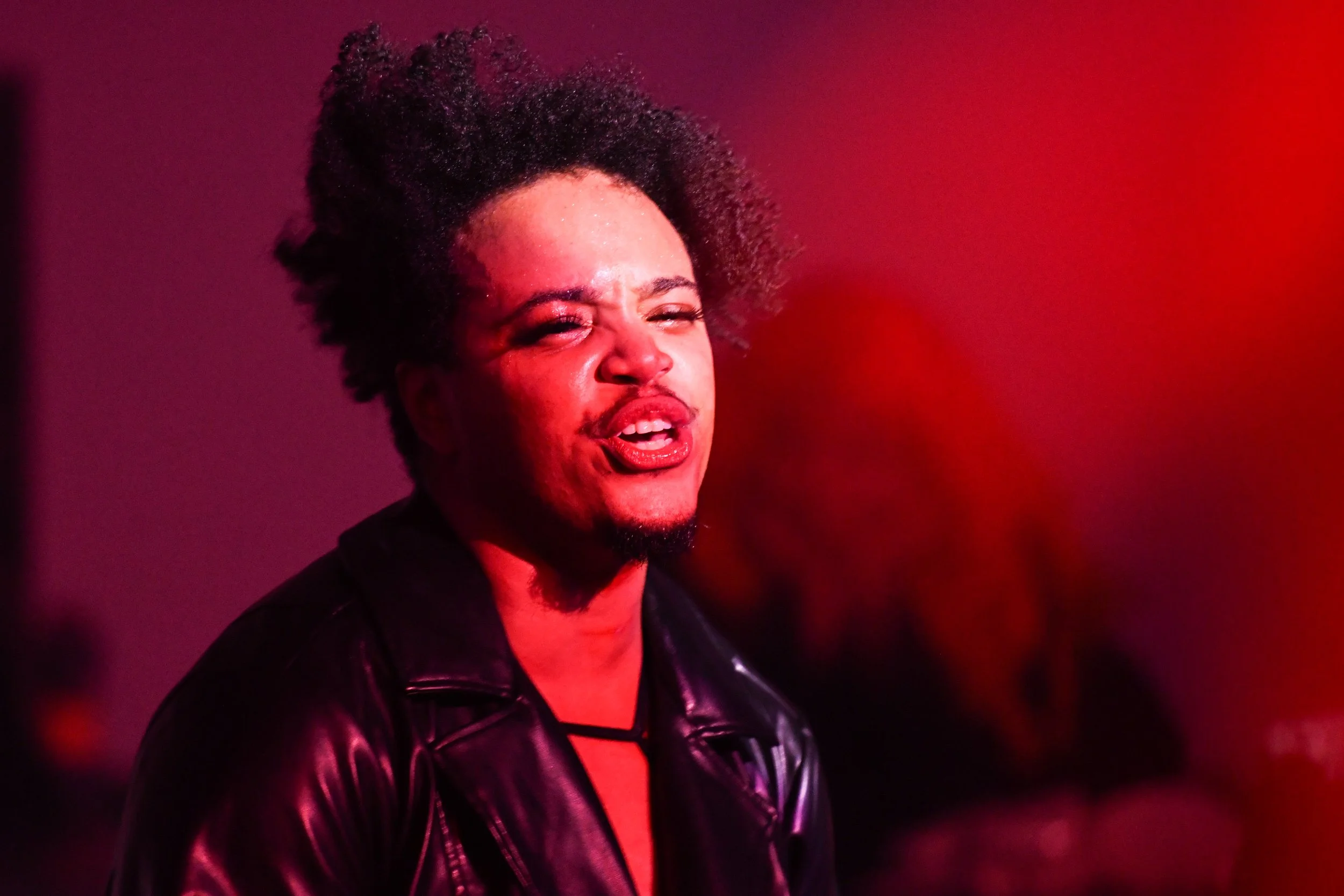 A person with curly hair and a goatee wearing a leather jacket, speaking against a red background.