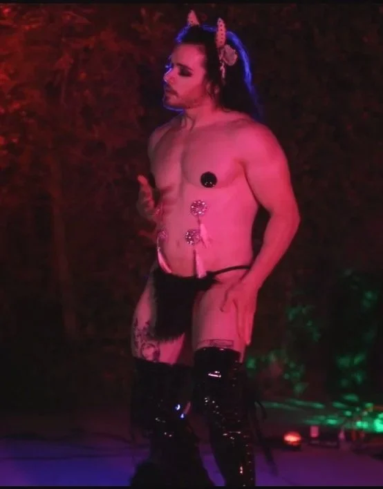 Person with blue hair wearing unicorn horn headband, topless with pasties, black shorts, and thigh-high black boots, standing in a dimly lit scene with colorful lighting.