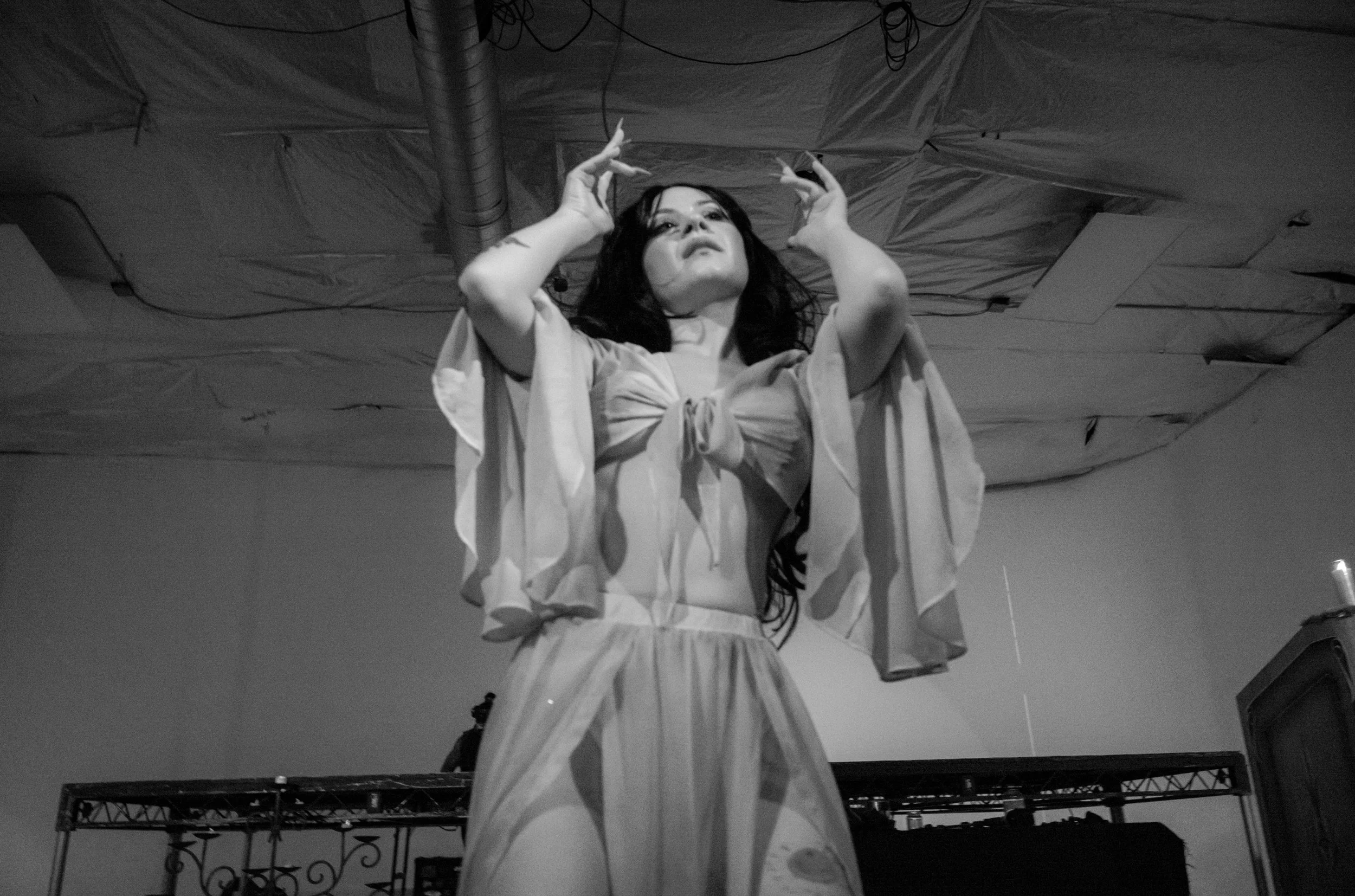 A woman with long dark hair wearing a light-colored dress, standing in front of a draped backdrop with industrial ducting above, holding her hands near her head.