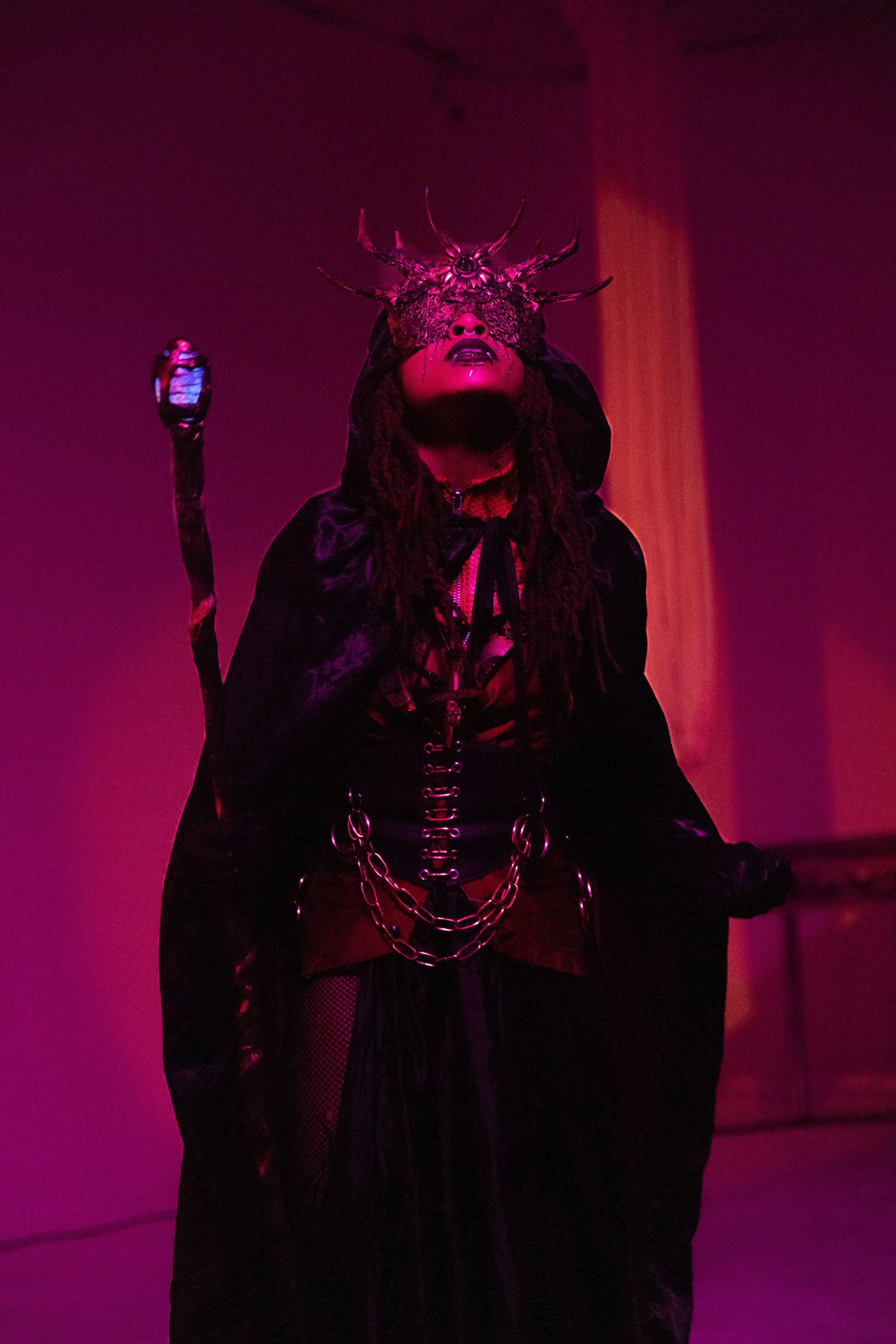Person dressed in dark, gothic costume with chains, wearing a lace mask and an elaborate headpiece with tendrils, in a dark, pink-lit environment.