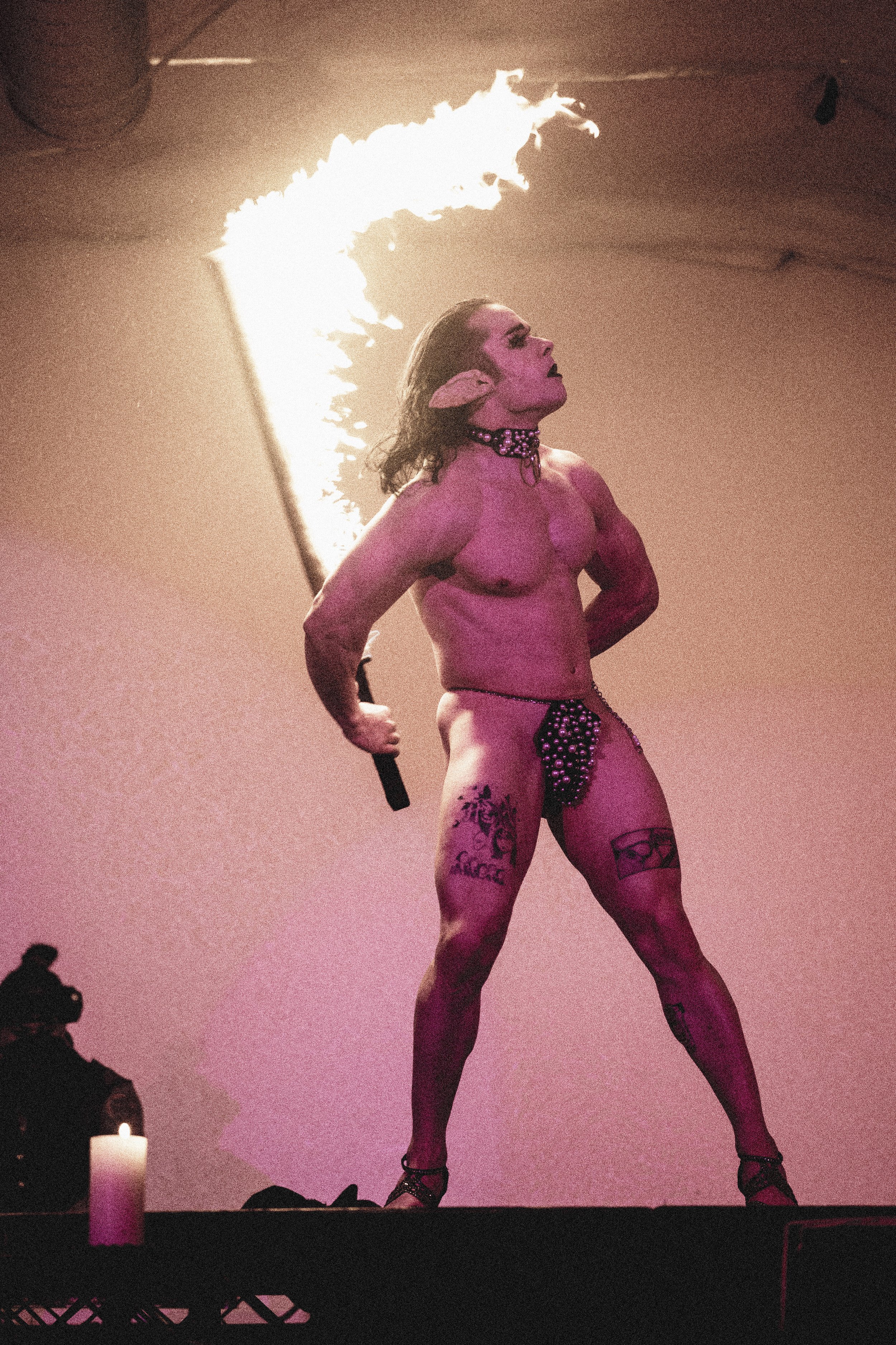 Performer with pink skin, tattoos, and elf ears stands on stage with a fire-breathing torch behind her. She wears minimal clothing, a choker, and high-heeled shoes, with her hands on her hips.