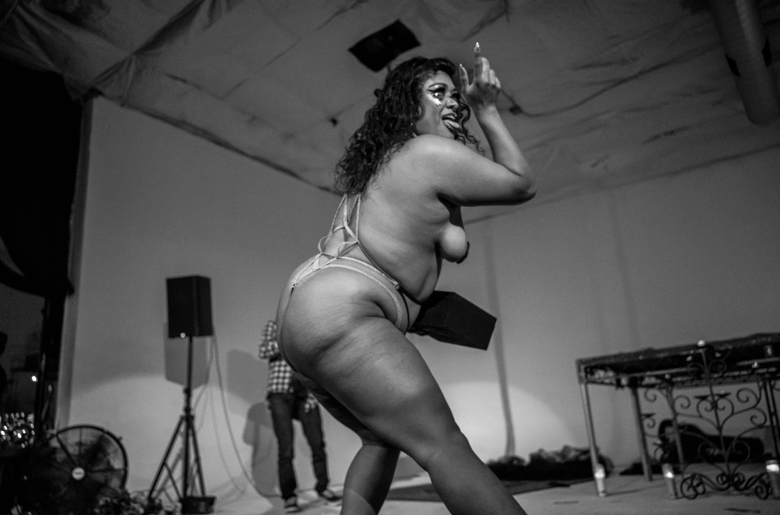 A woman with curly hair and tattoos is performing on stage in a dance pose, sticking out her tongue. She's wearing minimal clothing and appears to be dancing in front of a sound system and stage equipment.