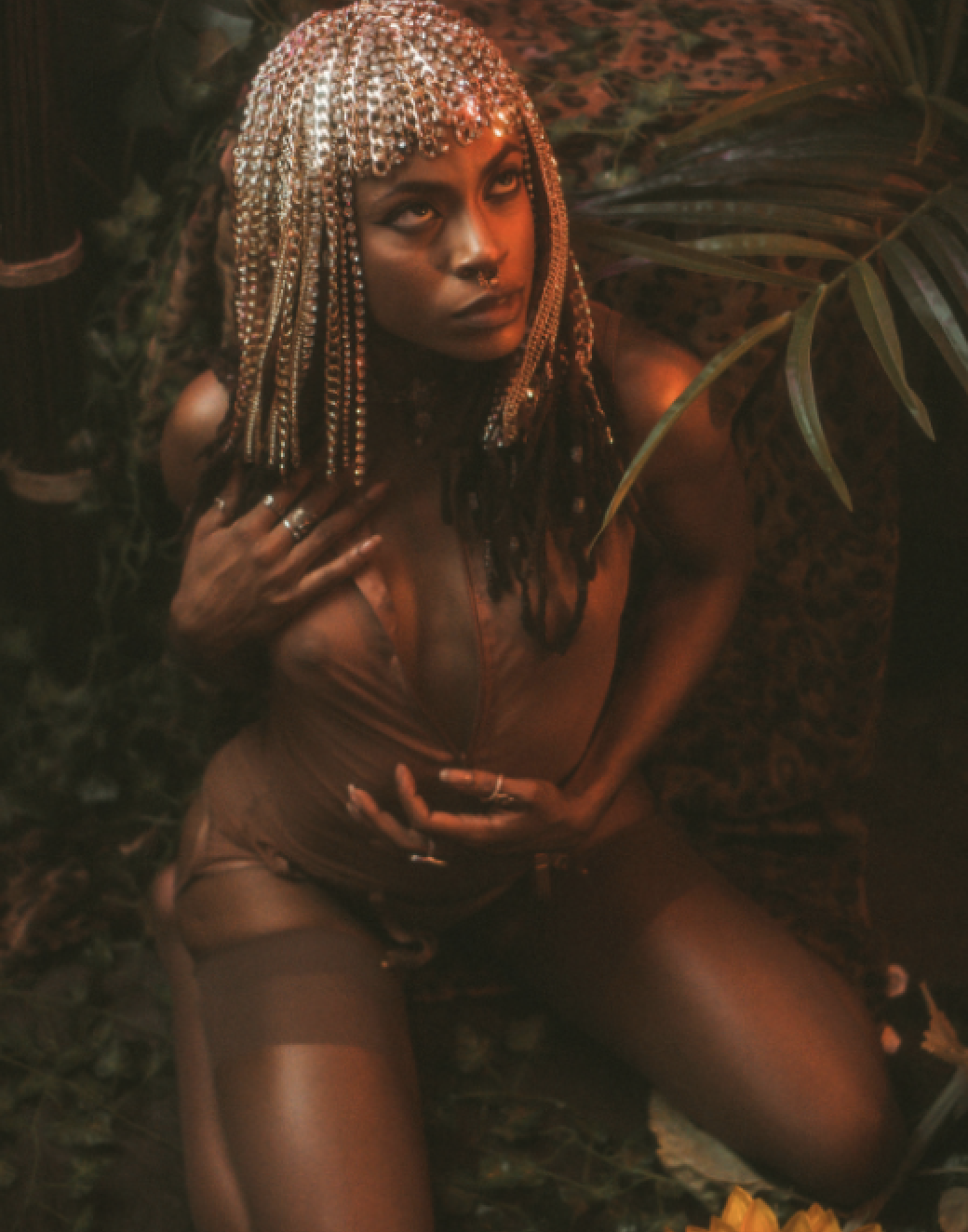 A woman with bead-embellished hair, wearing rings, posing among green plants in a dimly lit setting with a warm, reddish hue.