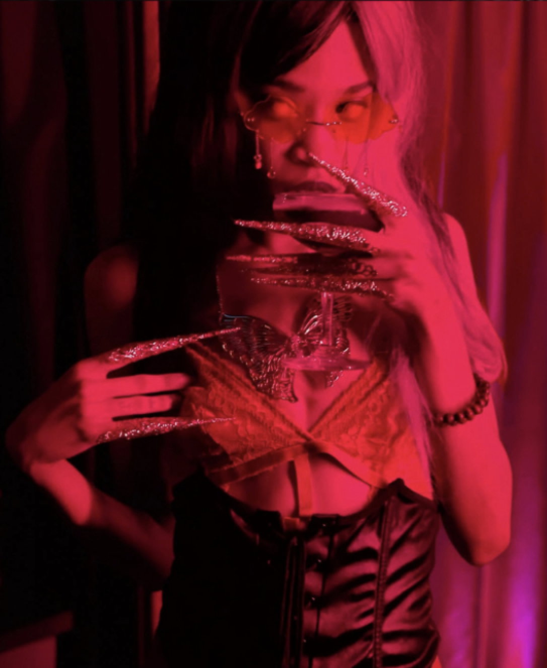 A woman dressed in dark, lace clothing is illuminated by red lighting. She is wearing unusual glasses and jewelry, posing with her hand near her mouth.