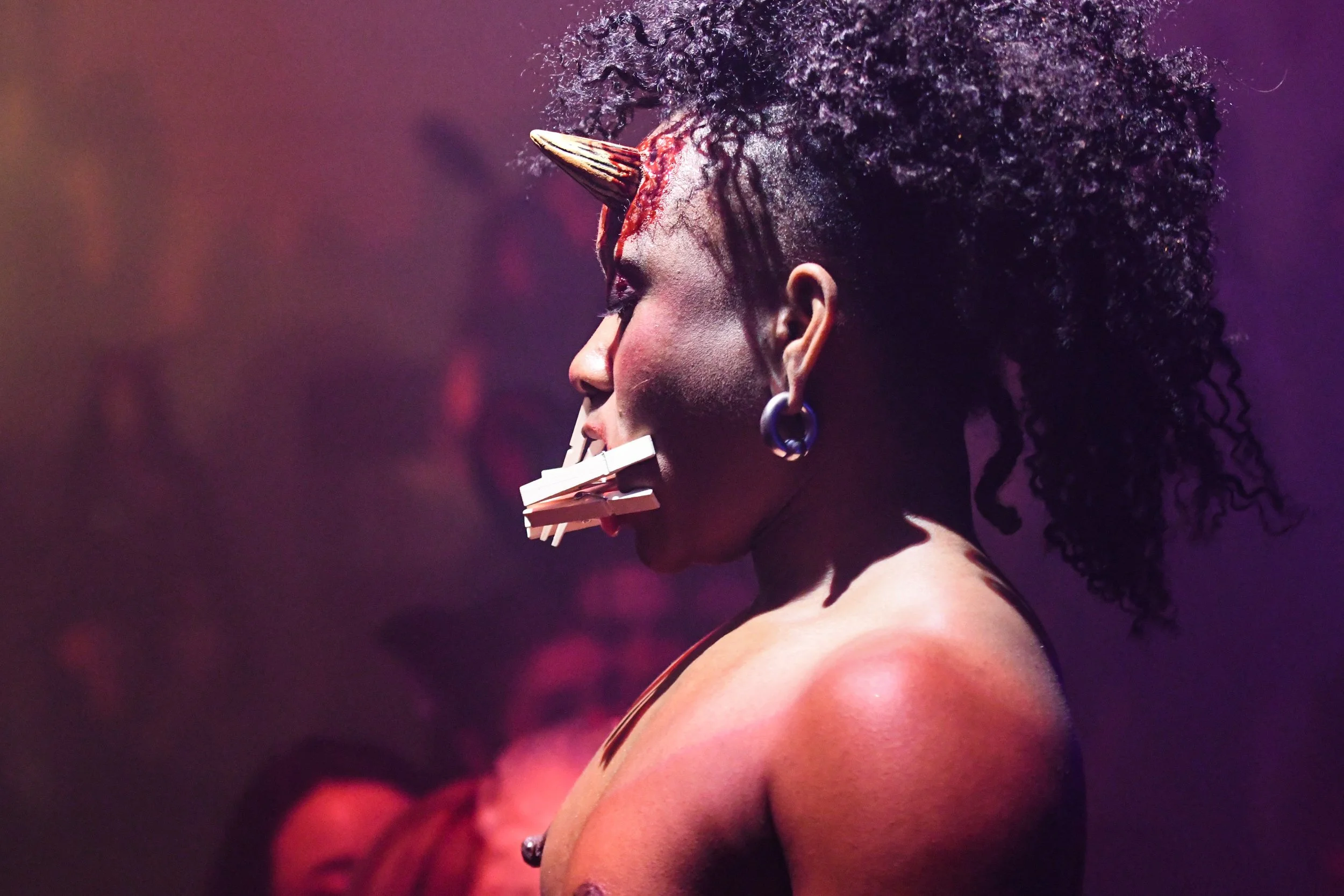 A woman with curly black hair, measuring tape around her neck, and a large pin/jewel on her forehead during a performance or event.