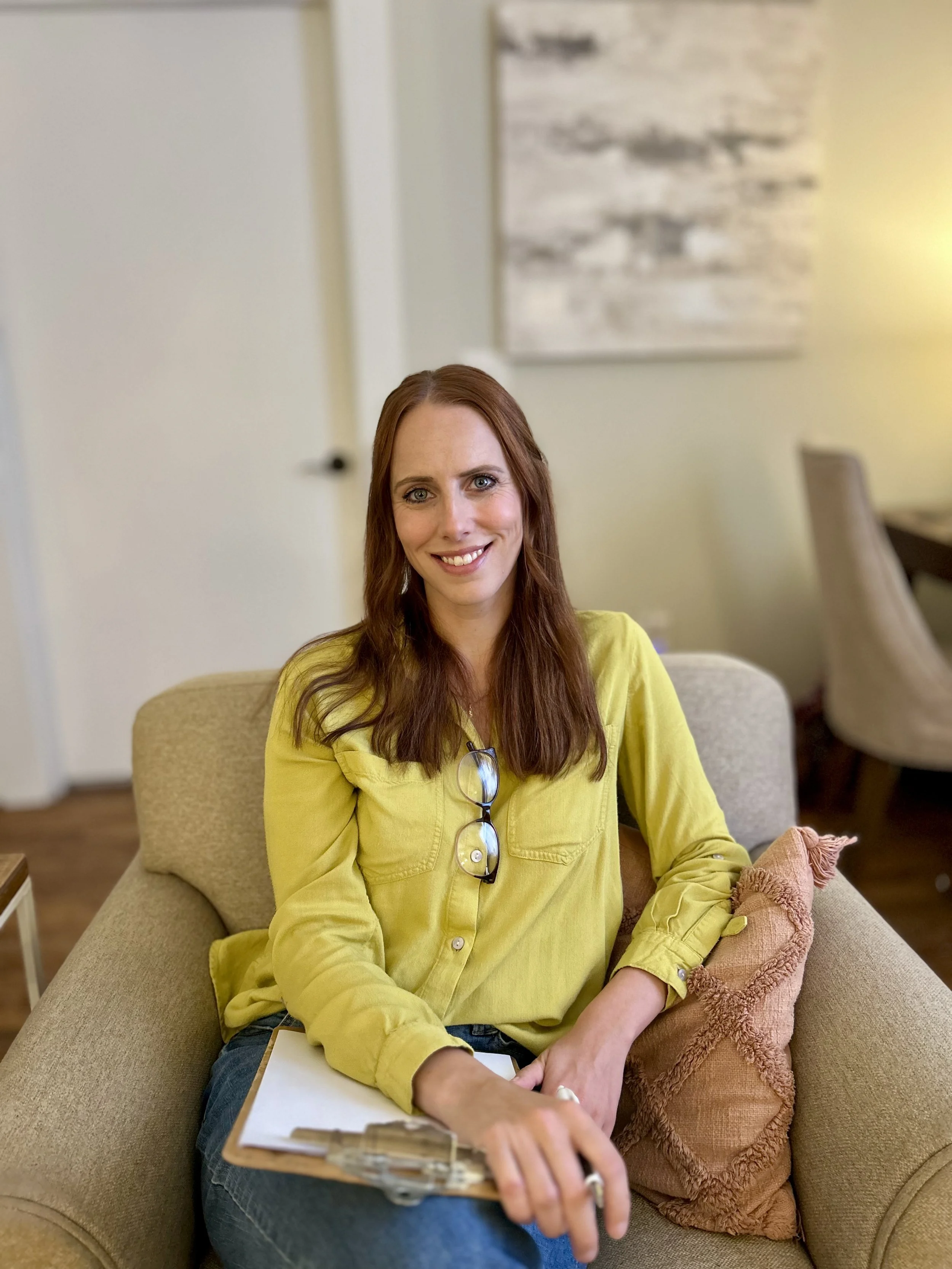 A woman with long red hair, wearing a yellow shirt, sitting on a beige sofa in a cozy room with a pillow, holding a clipboard and smiling at the camera.
