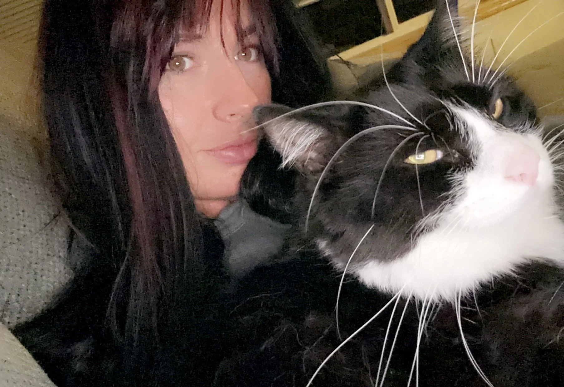 A woman with dark hair and light-colored eyes lying on a carpeted floor, close to a large black and white cat with yellow eyes.