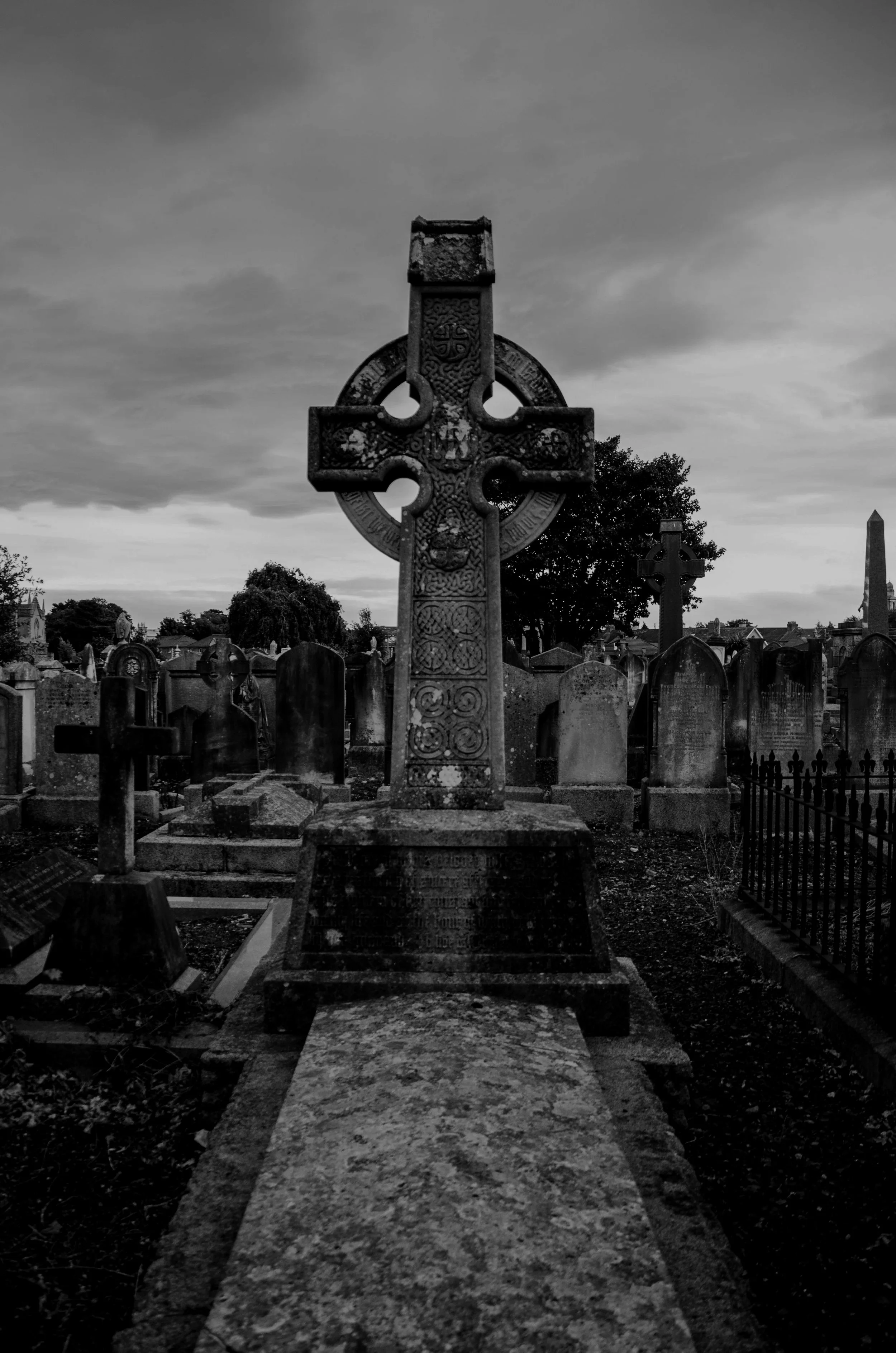 A black and white photo of a graveyard with a prominent Celtic cross headstone in the center, surrounded by other tombstones and a cloudy sky in the background.