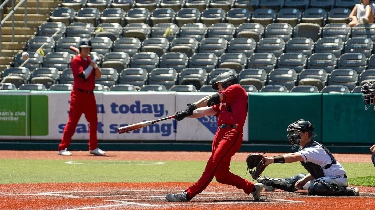 A baseball player in a red uniform swings a bat at a pitched ball, while the catcher in a black and white uniform kneels behind home plate. An umpire in the background watches, with empty stadium seats visible beyond.
