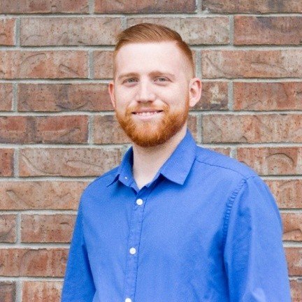 Owner and Fora Advisor Derek Rouse, a man with red hair and beard wearing a blue shirt. He is standing in front of a brick wall.
