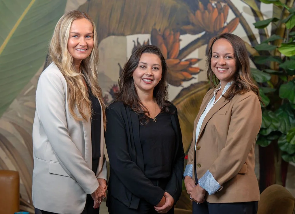 Three women standing together indoors in front of a tropical-themed mural, smiling at the camera, dressed in business casual attire.